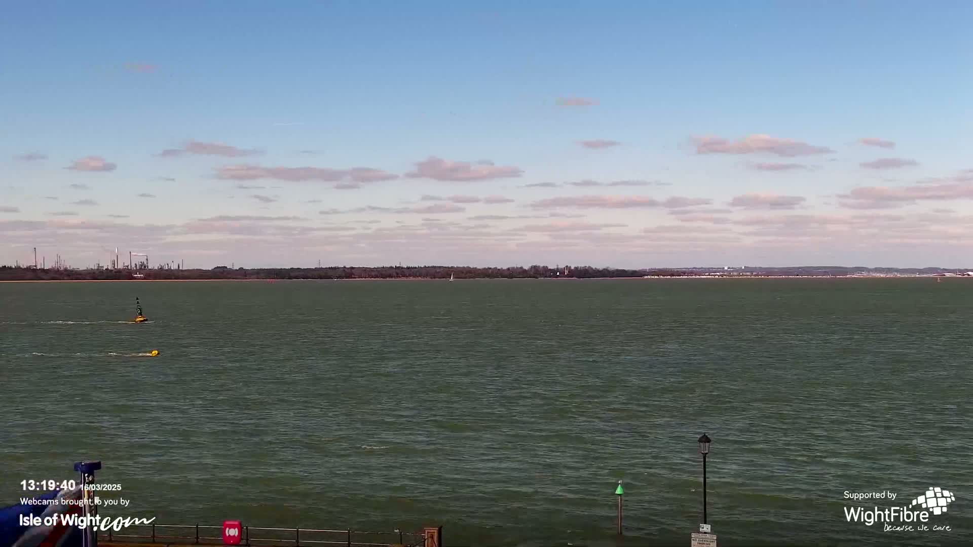 A calm body of water stretches to a distant shoreline under a partly cloudy sky, with two small objects floating on the water's surface and an industrial area visible in the background.