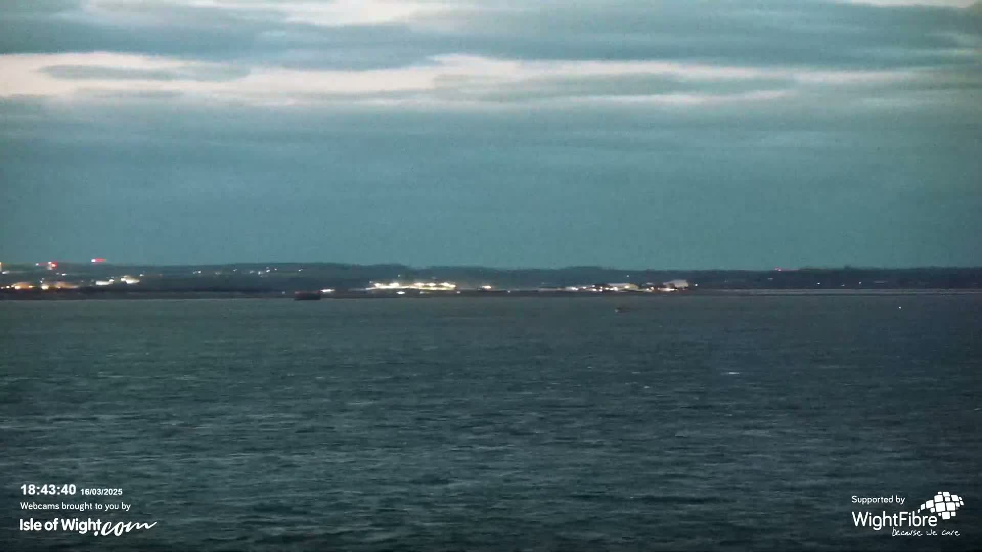 A dark, overcast sky hangs over a calm body of water, with a distant shoreline faintly illuminated at twilight.