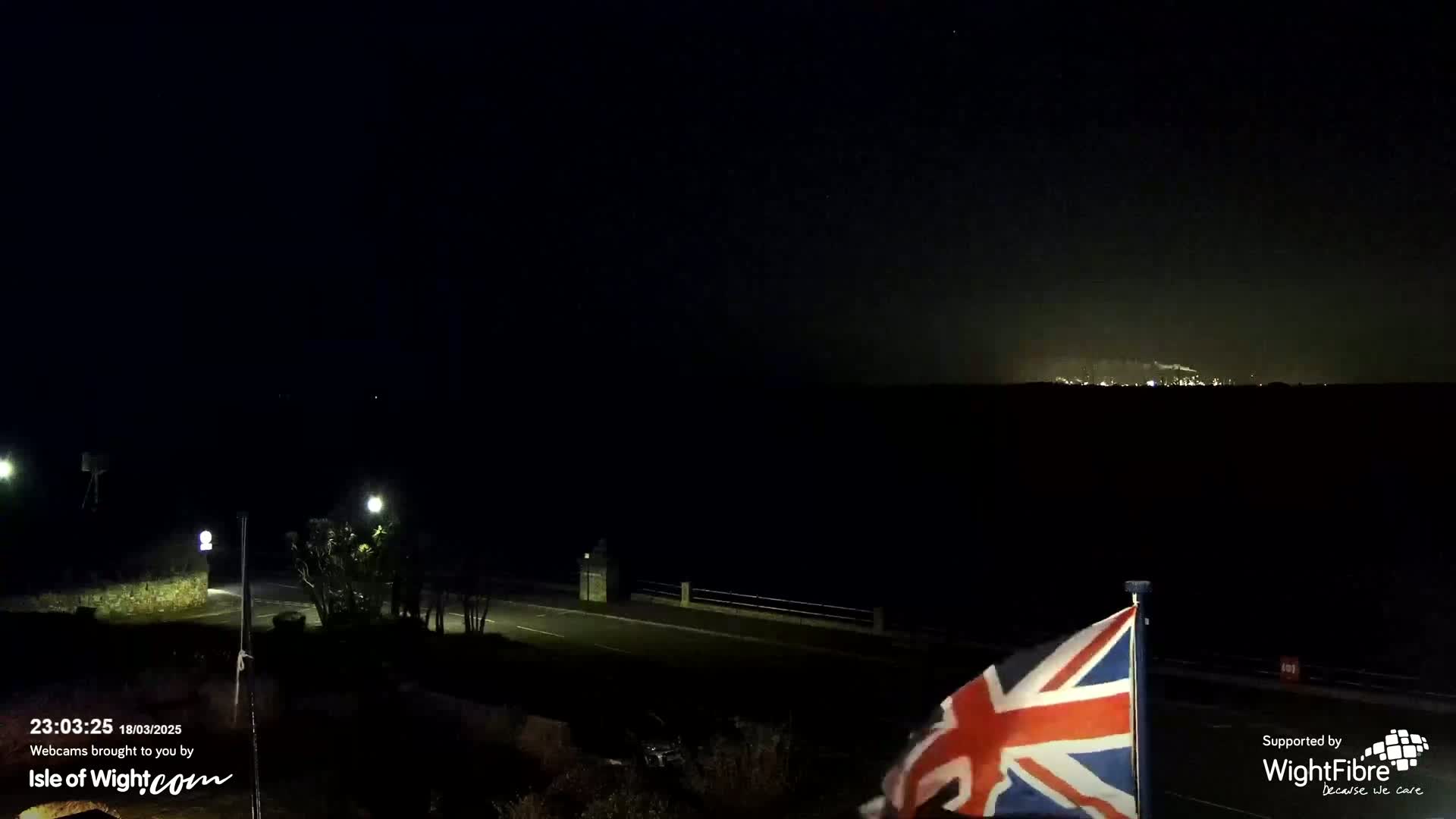 A nighttime view of a dark coastline with a faintly lit distant landscape under a clear, dark sky, and a Union Jack flag waving gently in the foreground.