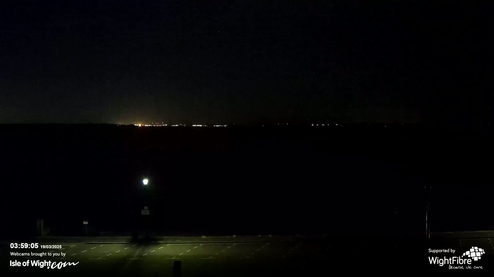 A dark, empty parking lot is shown at night, with a distant line of lights visible on the horizon under a clear, dark sky.
