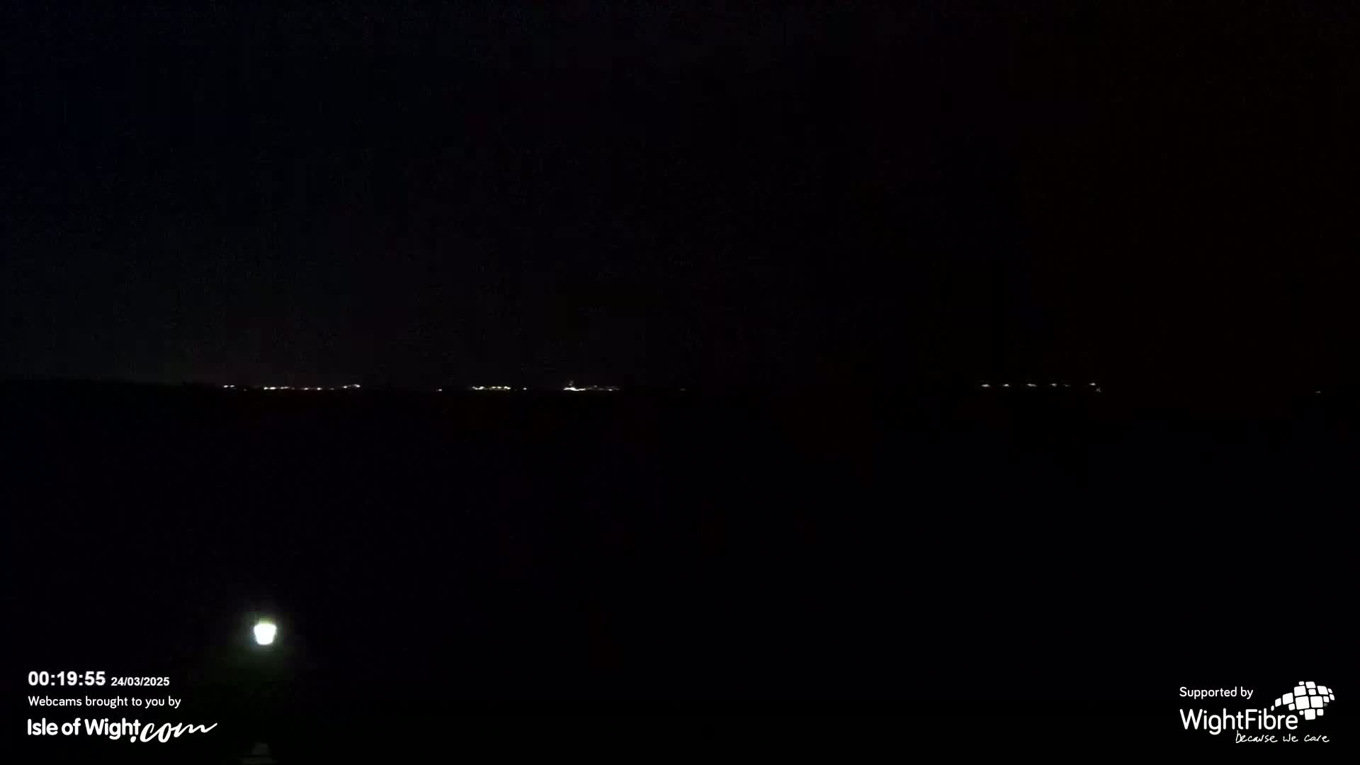 A nighttime, dark scene shows a distant line of faint lights on the horizon, illuminated by a bright light source in the lower left corner.