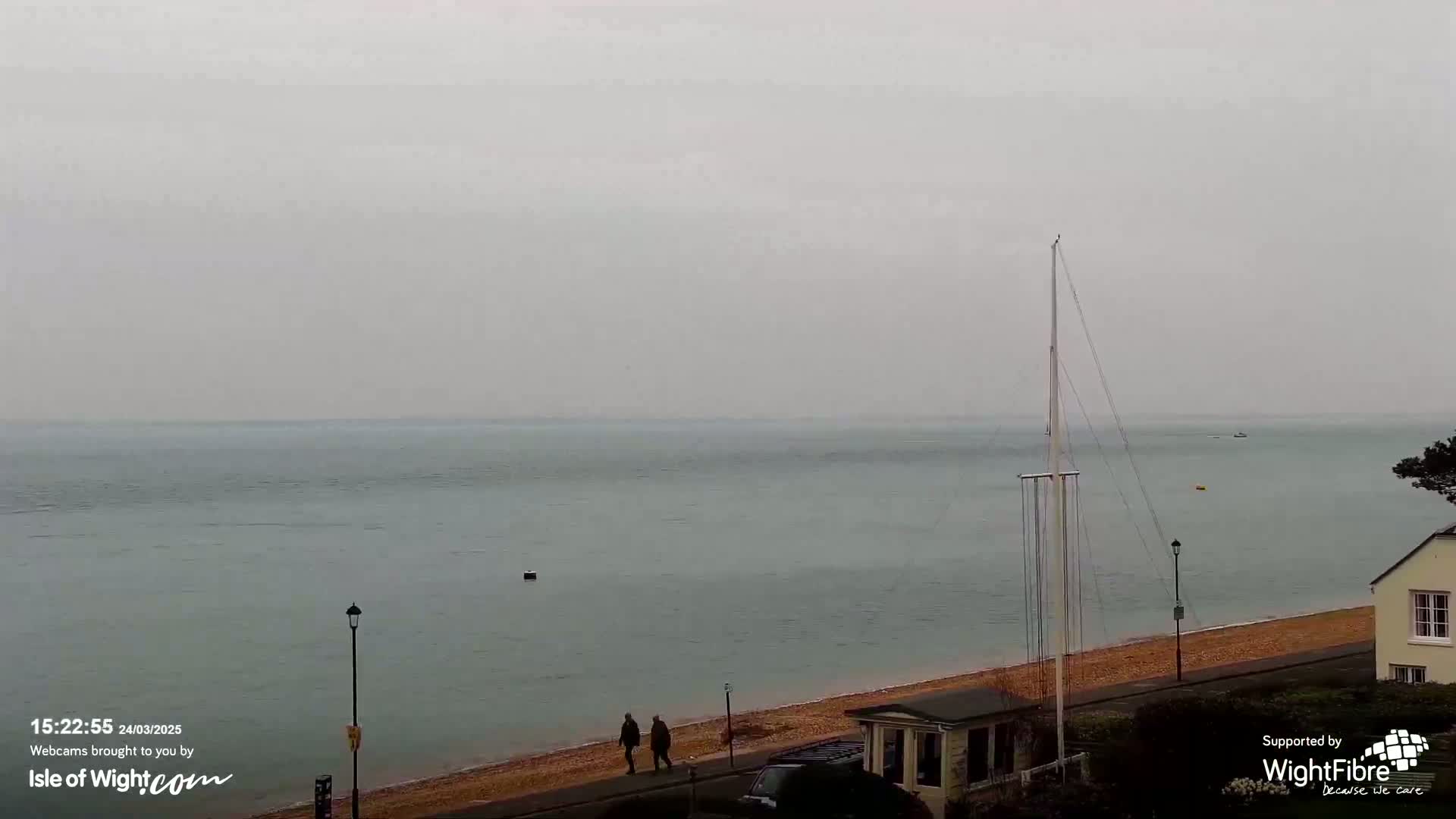 Two people walk along a beach next to calm, gray water under an overcast sky.