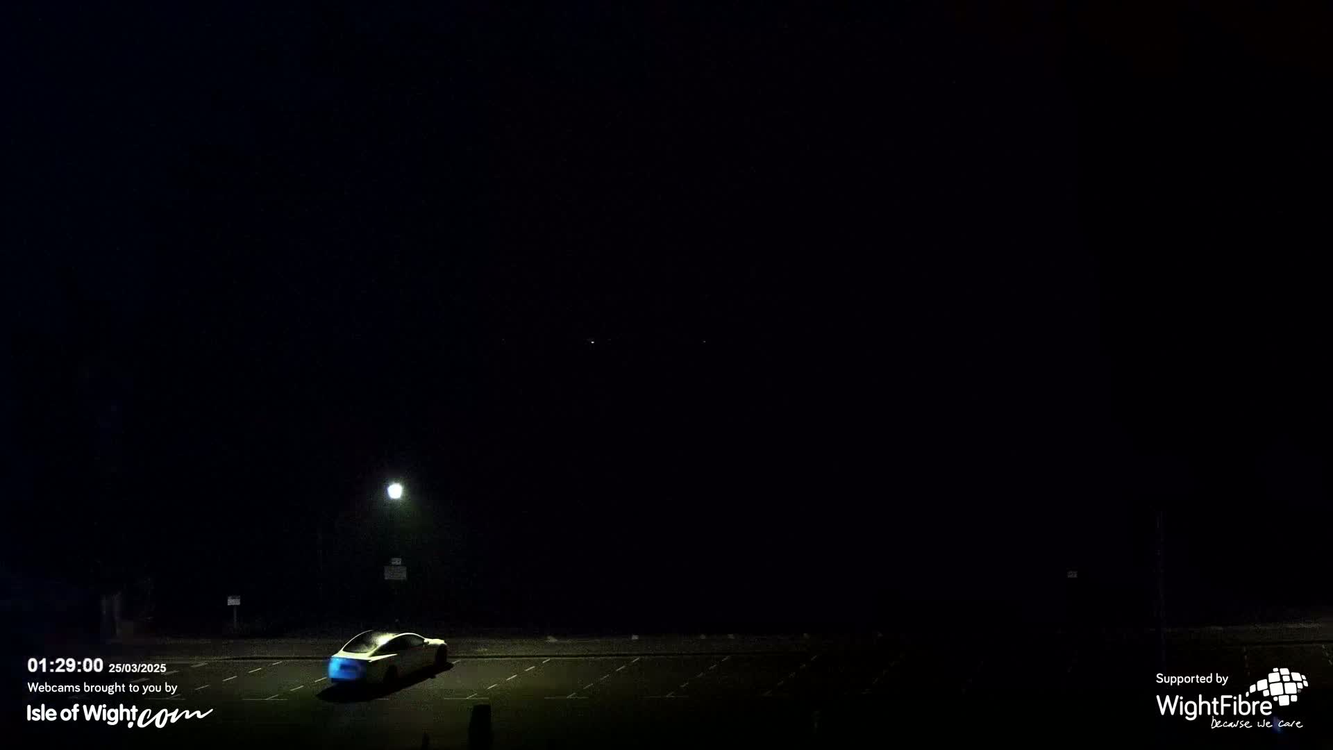 A single car sits in a dark, empty parking lot at night under a clear, dark sky illuminated only by a single lamppost.