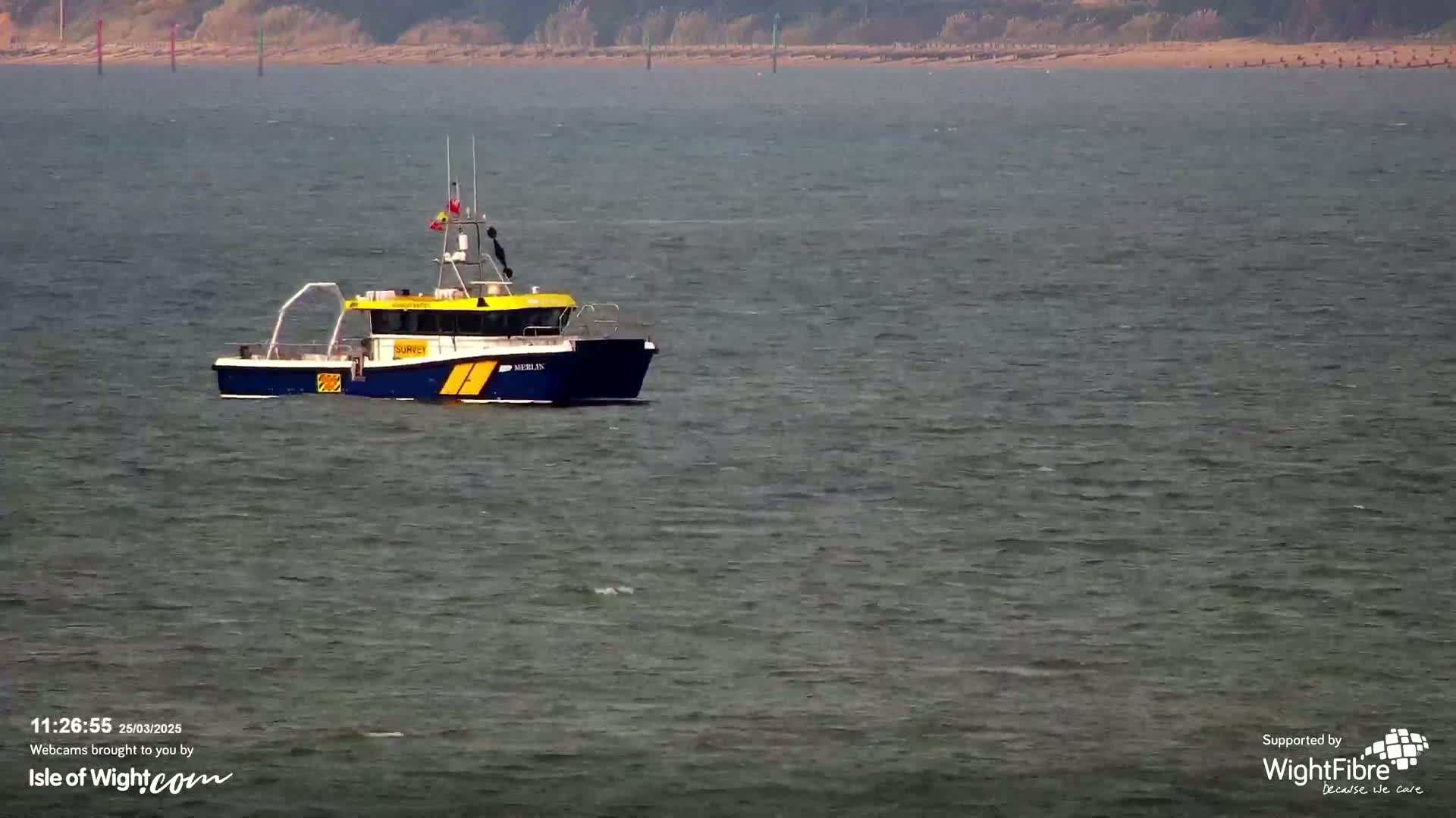 A small, blue and yellow survey boat floats on a calm, gray body of water with a distant shoreline visible.