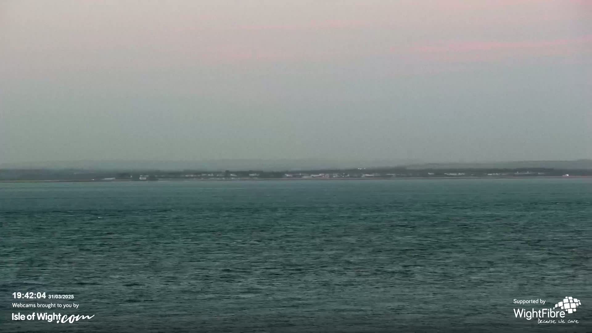 A hazy, overcast day shows a calm body of water with a distant shoreline featuring low-lying buildings and vegetation.