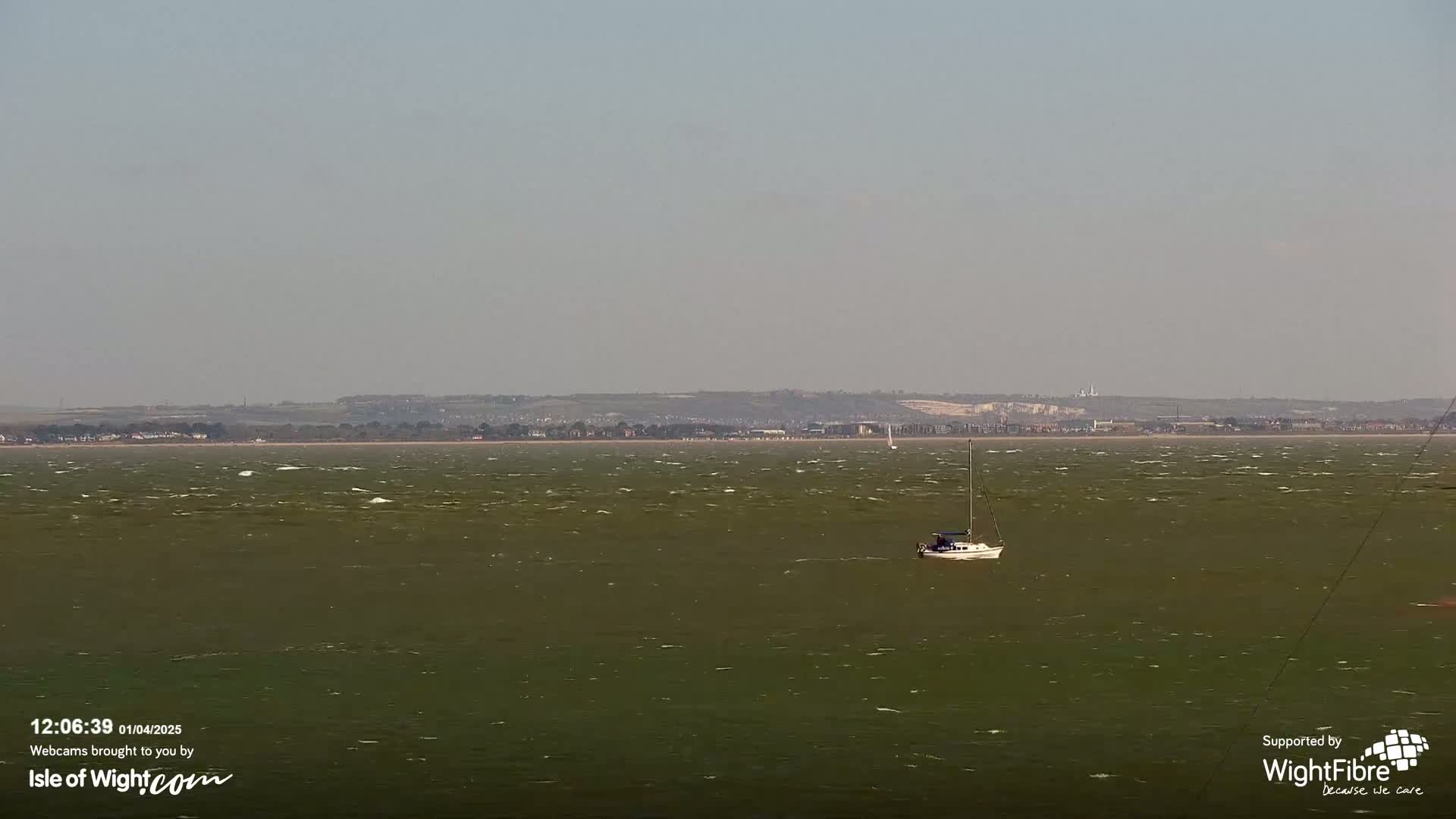 A sailboat floats on a choppy, greenish-grey body of water, with a distant shoreline visible under a hazy sky.