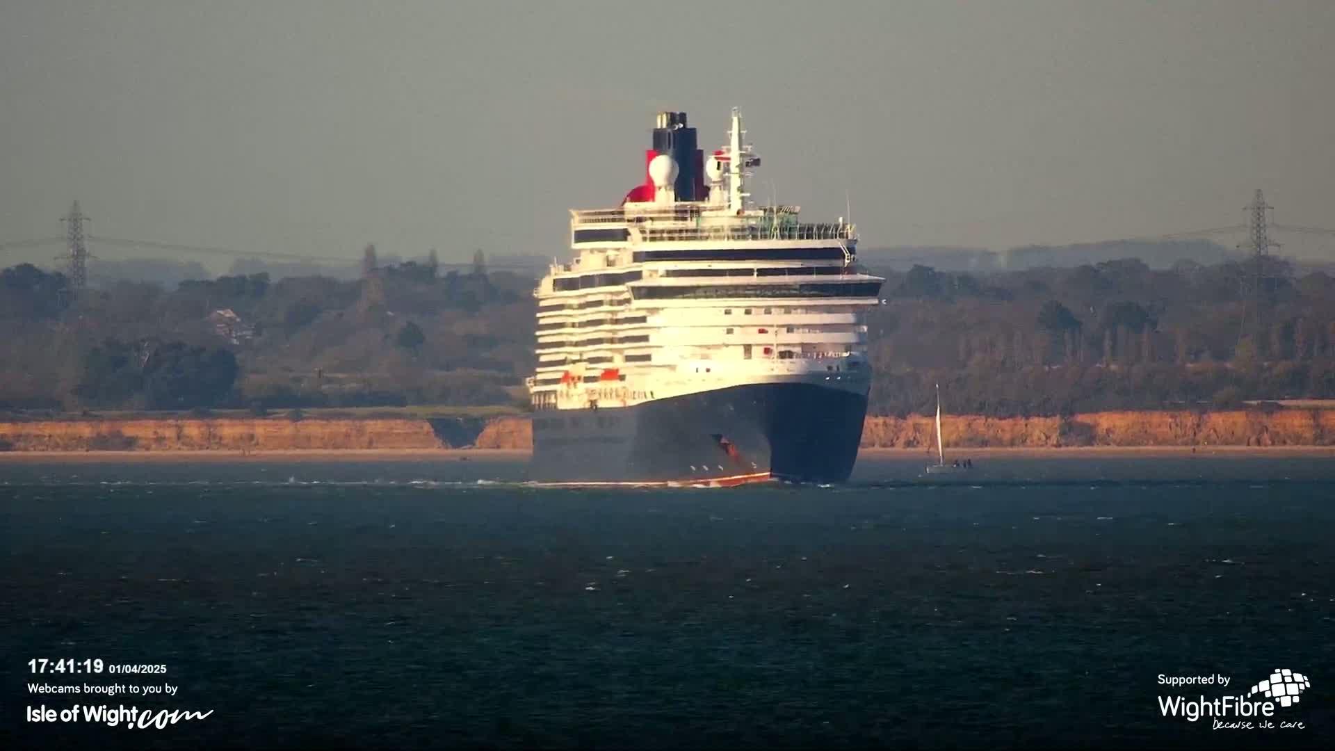 A large cruise ship sails on calm water past a shoreline with low, tan cliffs and a distant, hazy landscape.