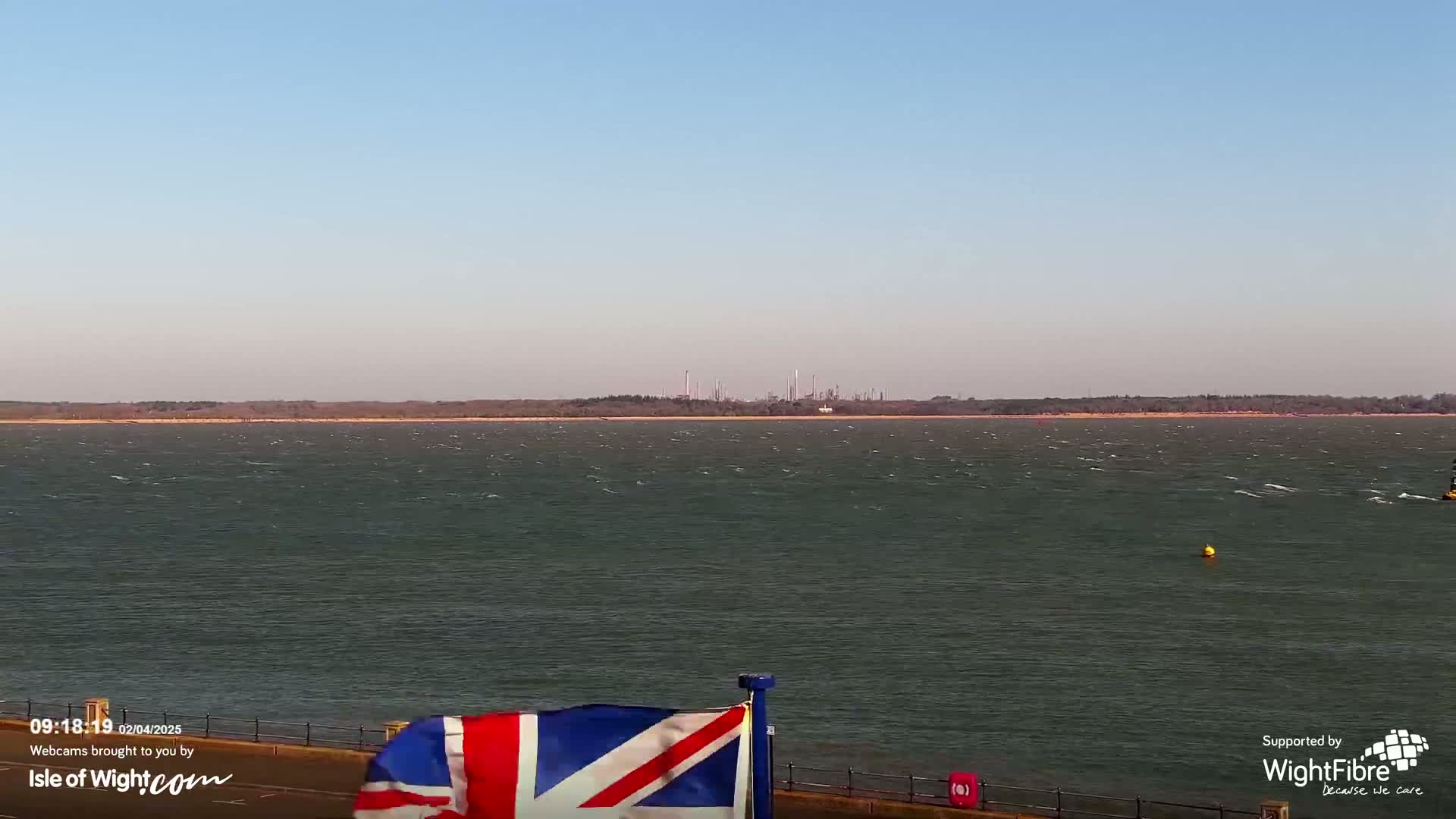 A body of water with small waves under a clear sky shows a distant industrial area on the horizon, and a Union Jack flag is partially visible in the foreground.