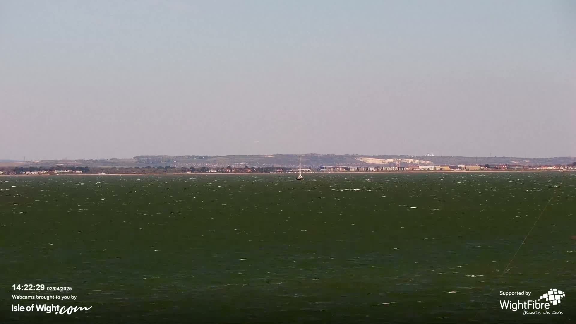 A sailboat sits in a dark-green body of water under a hazy sky with a distant coastline visible in the background.
