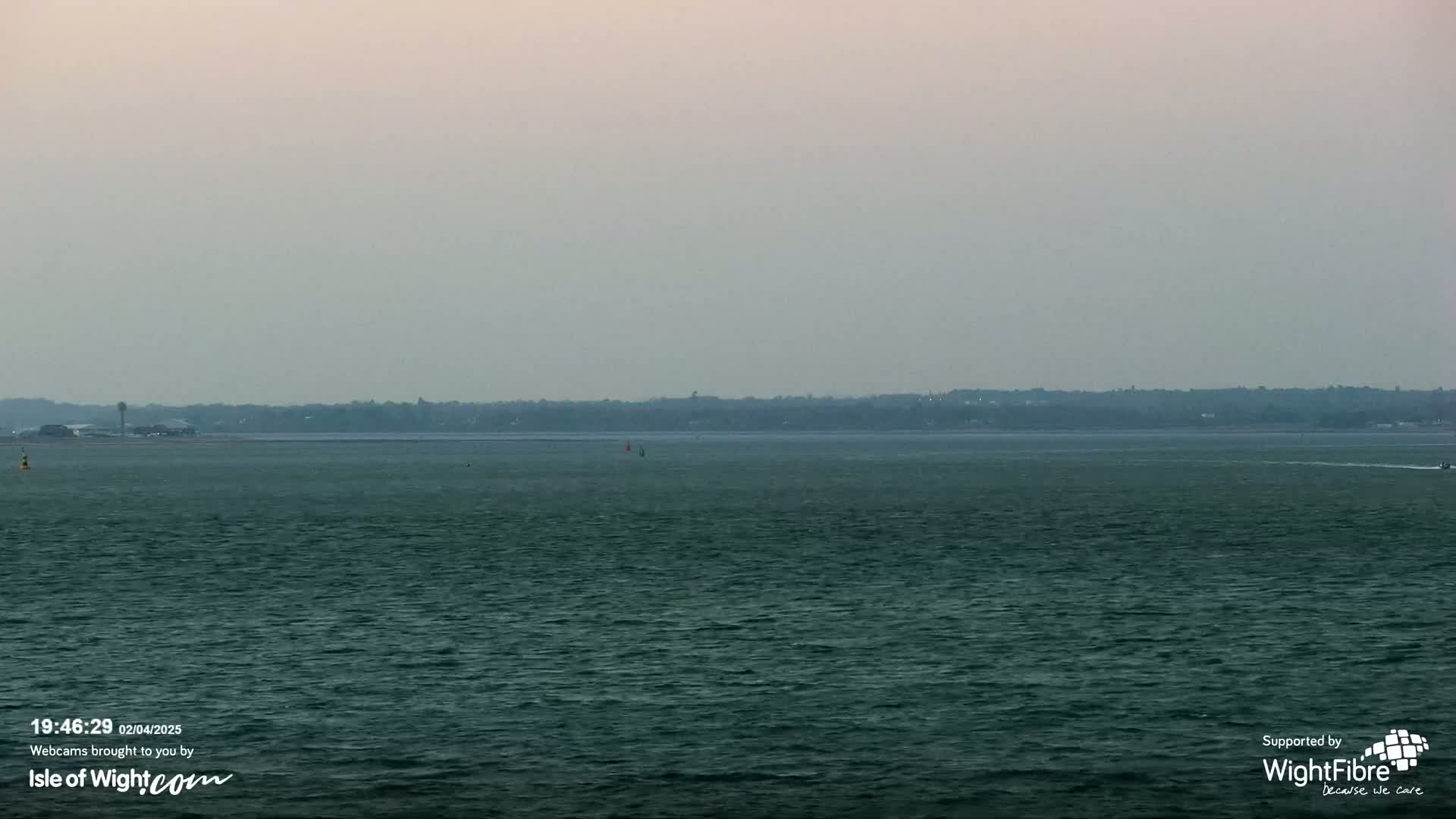 A hazy view of a calm, dark-green body of water shows a distant shoreline under a pale, overcast sky.