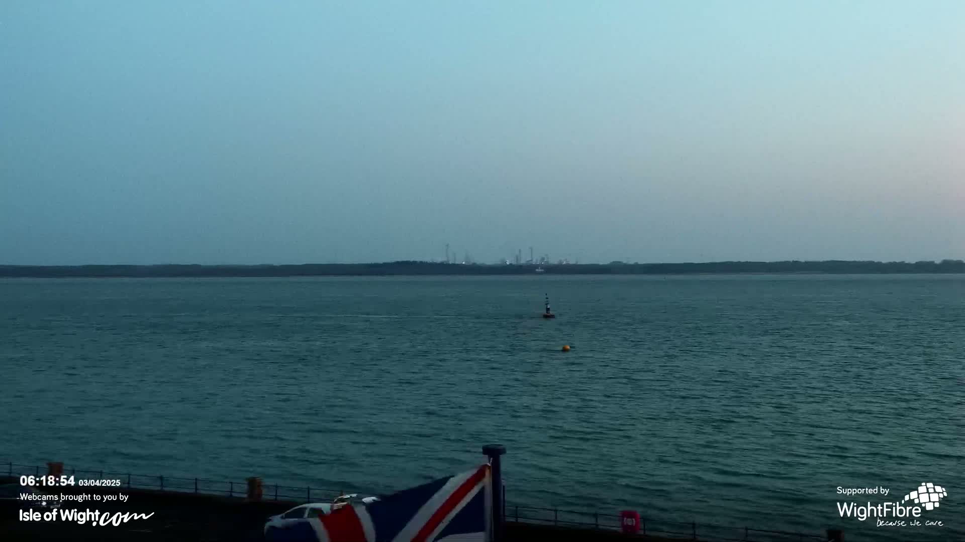 A calm, dark-blue body of water extends to a distant shoreline featuring an industrial complex under a hazy, light-blue sky.