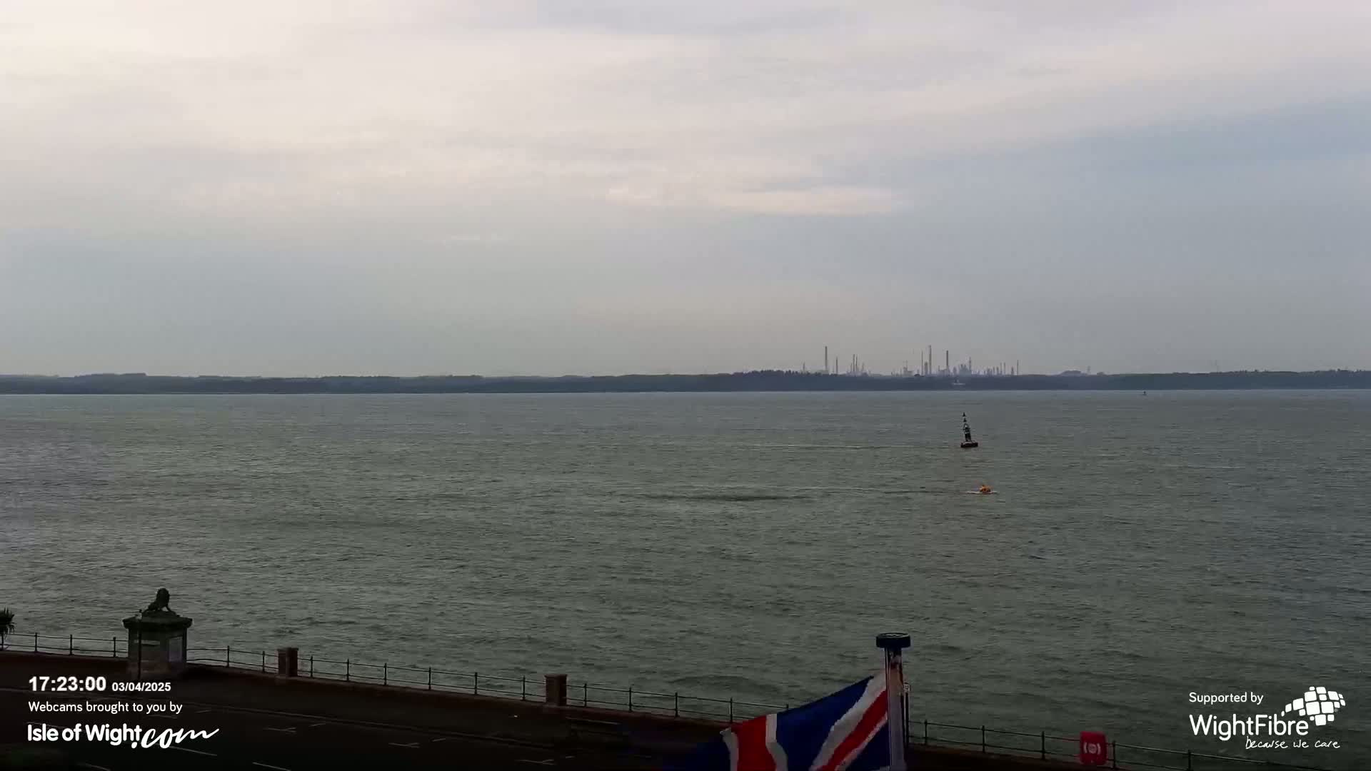 A calm, gray day shows a wide expanse of water with a distant industrial skyline and a small buoy in the foreground.