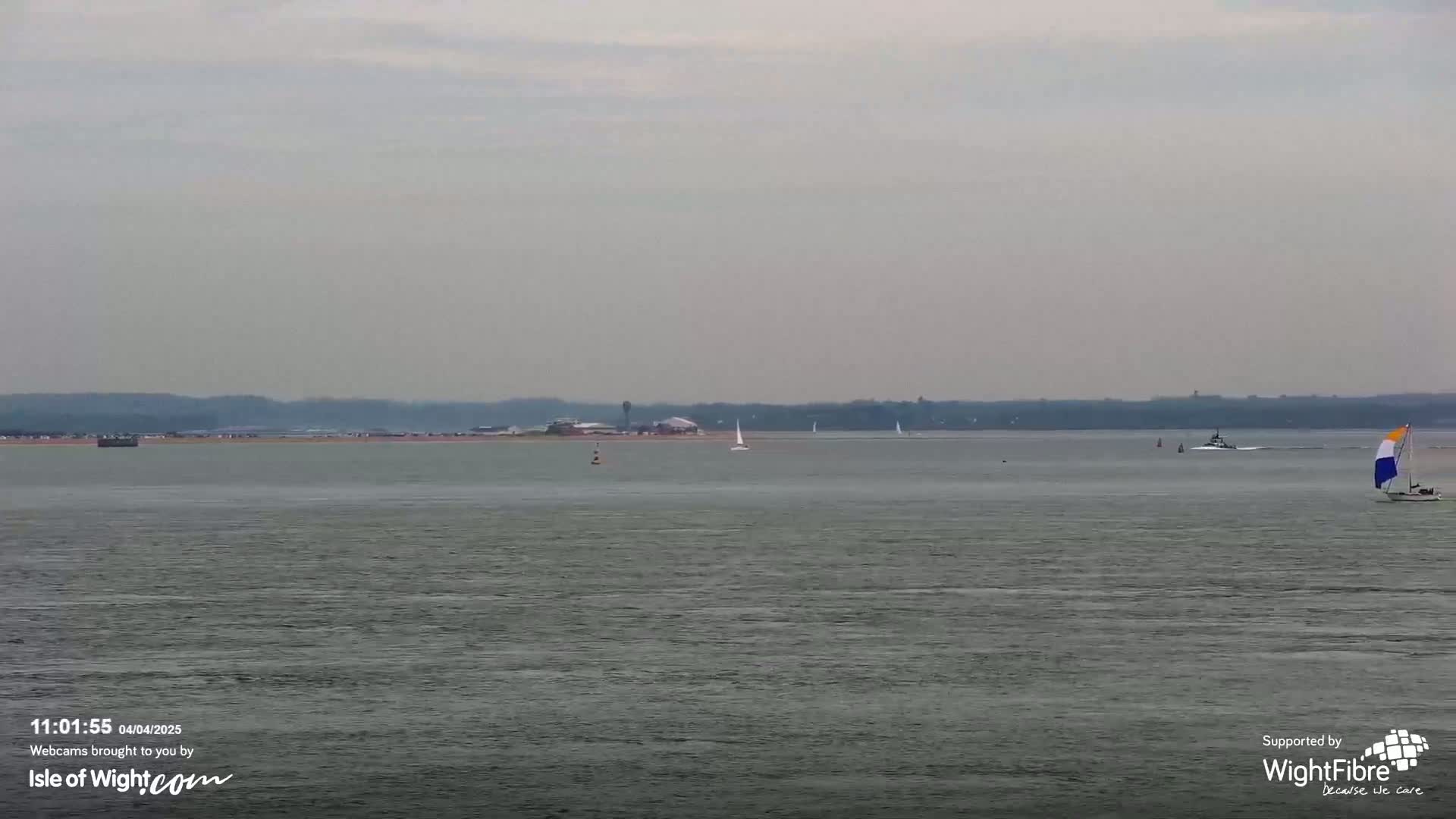 A calm, gray day shows a body of water with several sailboats and a small motorboat; in the distance, land with some buildings is visible.