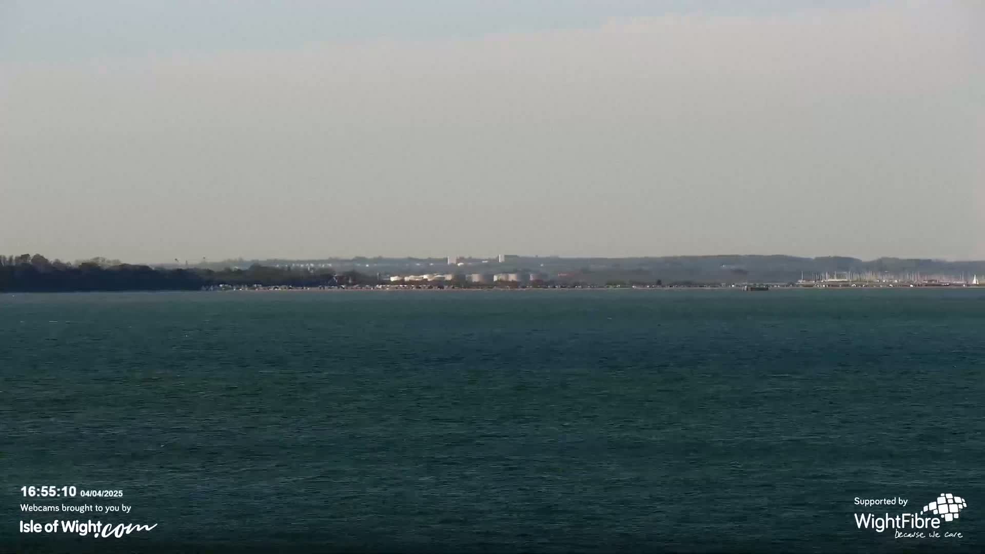 A calm body of water stretches to a distant shoreline, where land, buildings, and a beach with people are visible under a hazy sky.