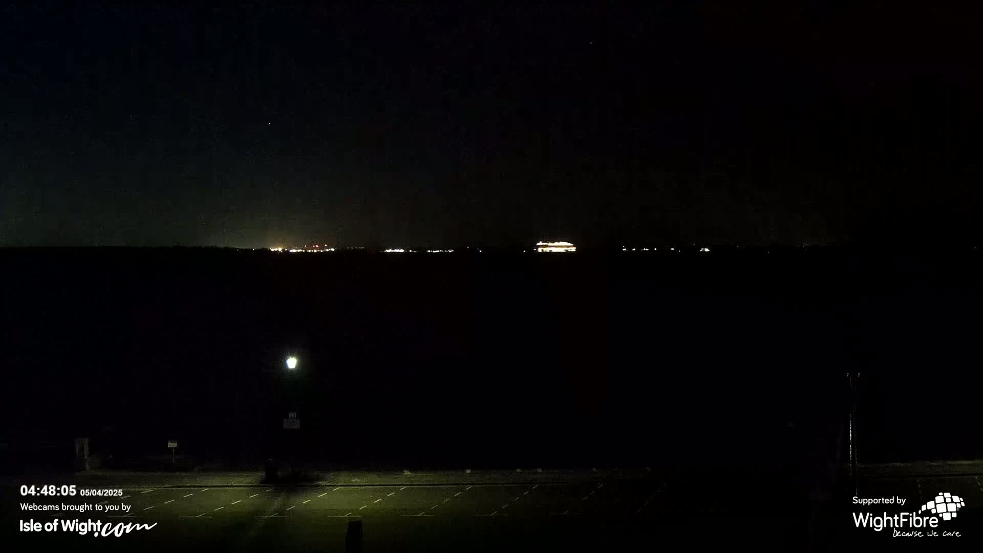 A dark, nighttime scene shows a distant, faintly lit horizon with a mostly empty, dark parking lot in the foreground under a clear, starry sky.