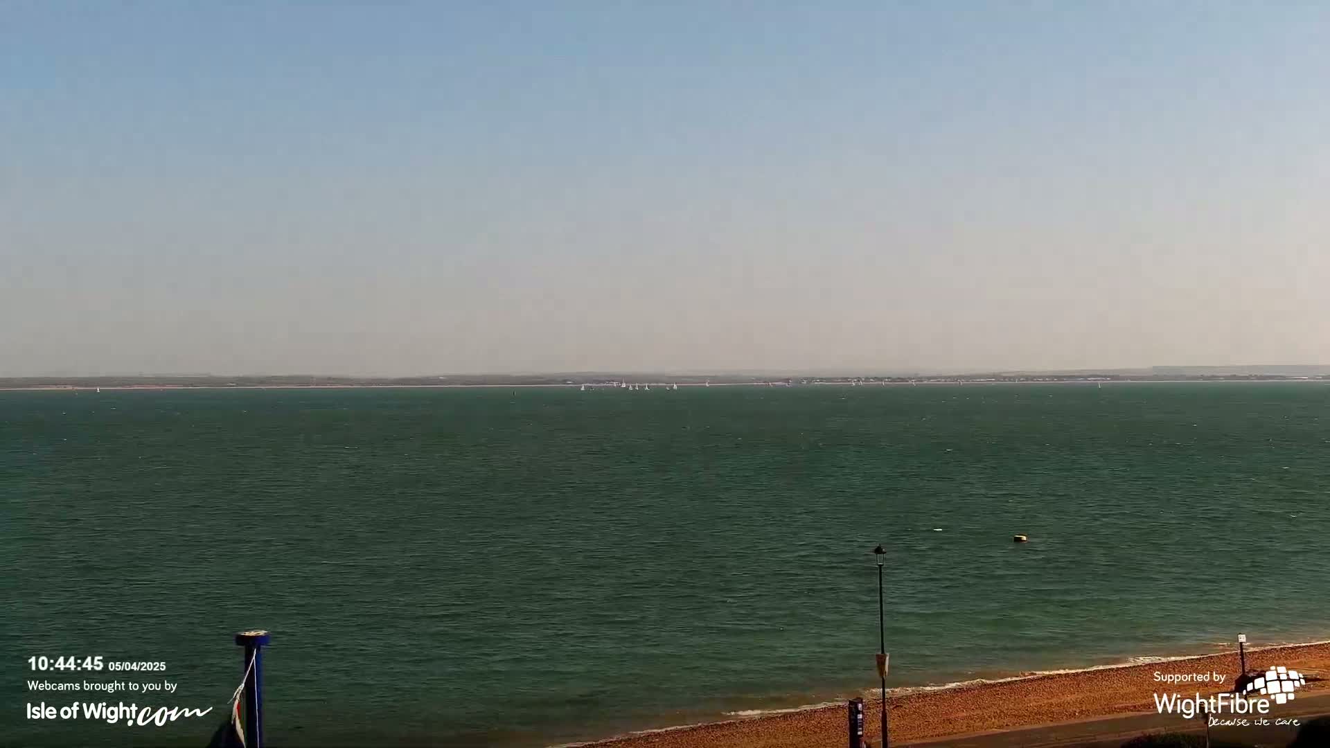 A calm, expansive body of water with several sailboats in the distance is viewed from a beach on a clear, sunny day.