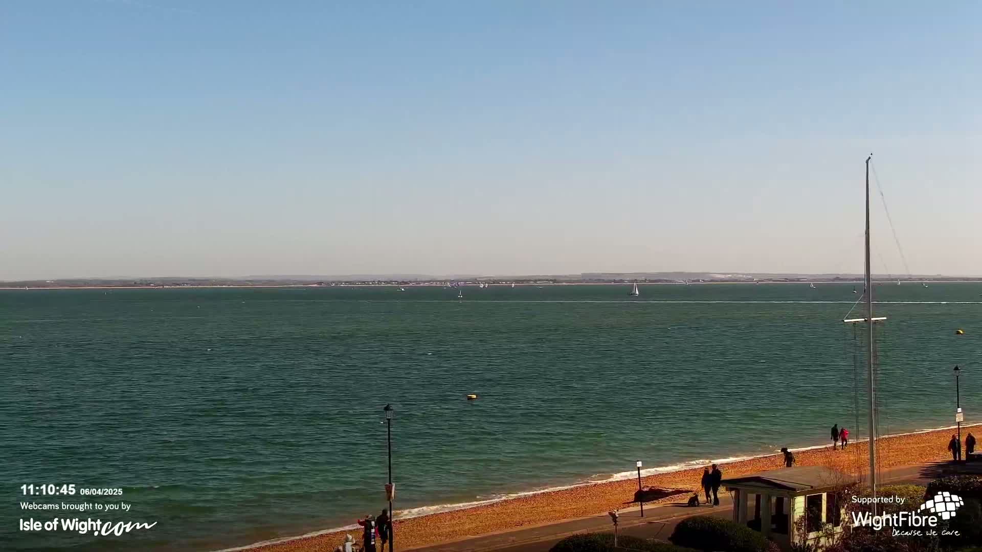 A sunny day shows a wide expanse of calm, dark-green water with several sailboats in the distance, a sandy beach with people strolling along the shore, and a tall mast on the right.