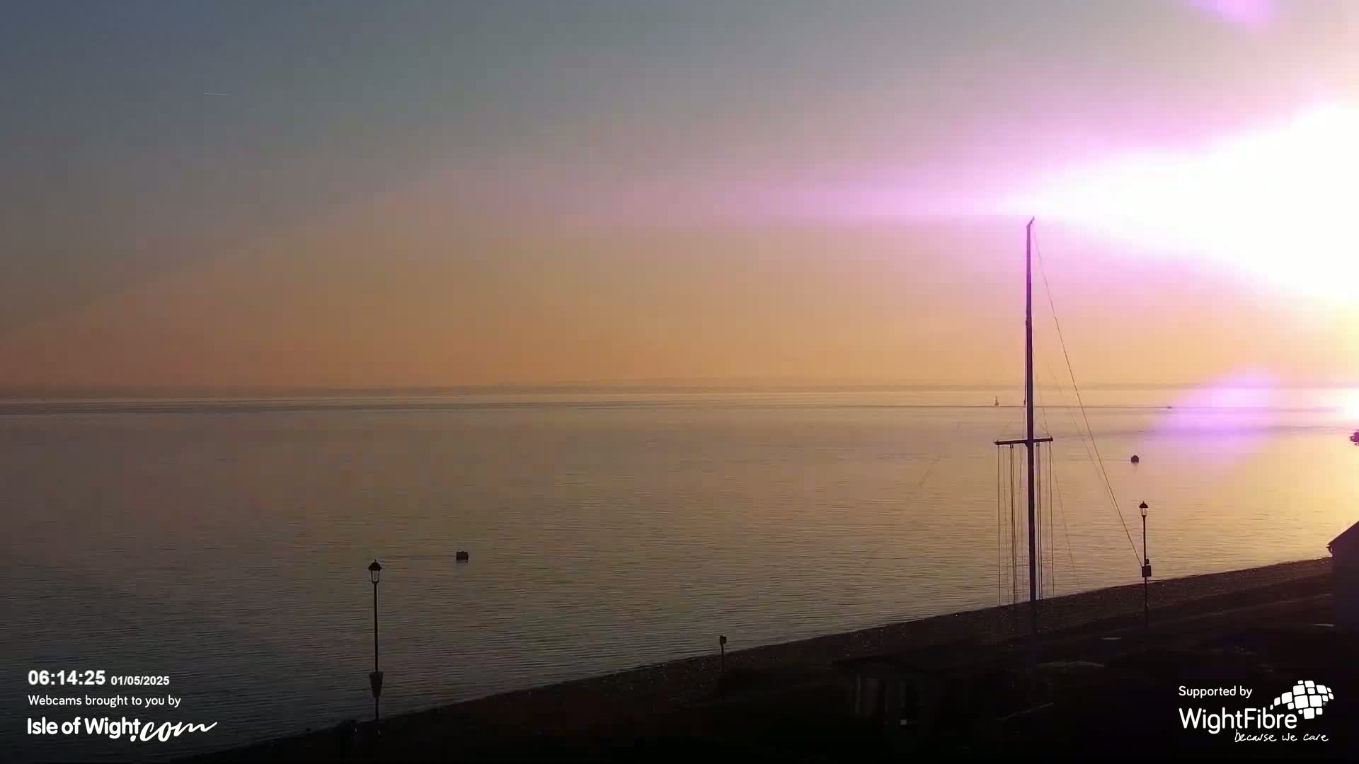 A calm sea at sunrise with a tall mast on the shore, under a pinkish-orange sky.