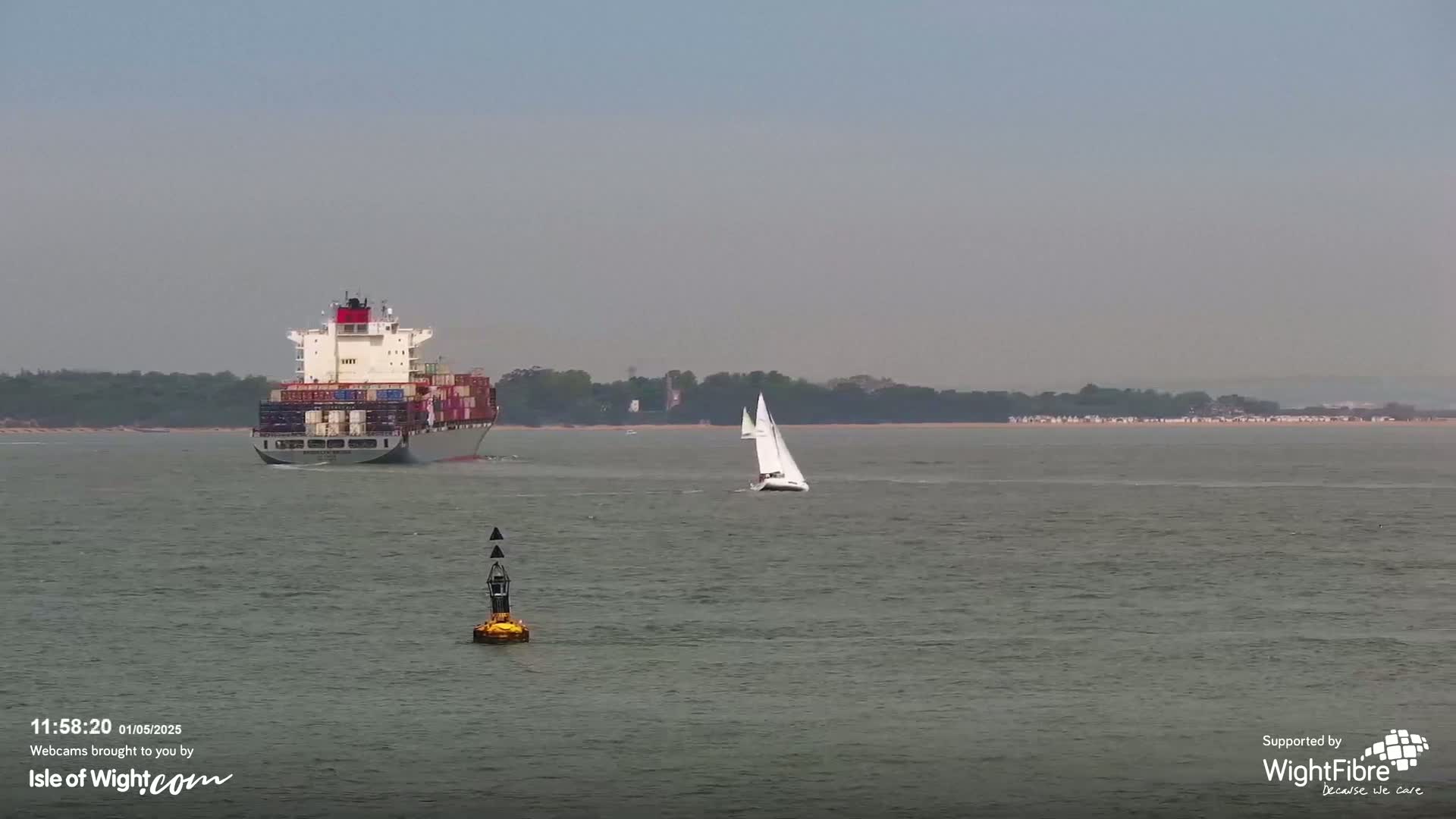 A large container ship and a sailboat are visible on a calm, gray body of water, with a distant shoreline in the background.