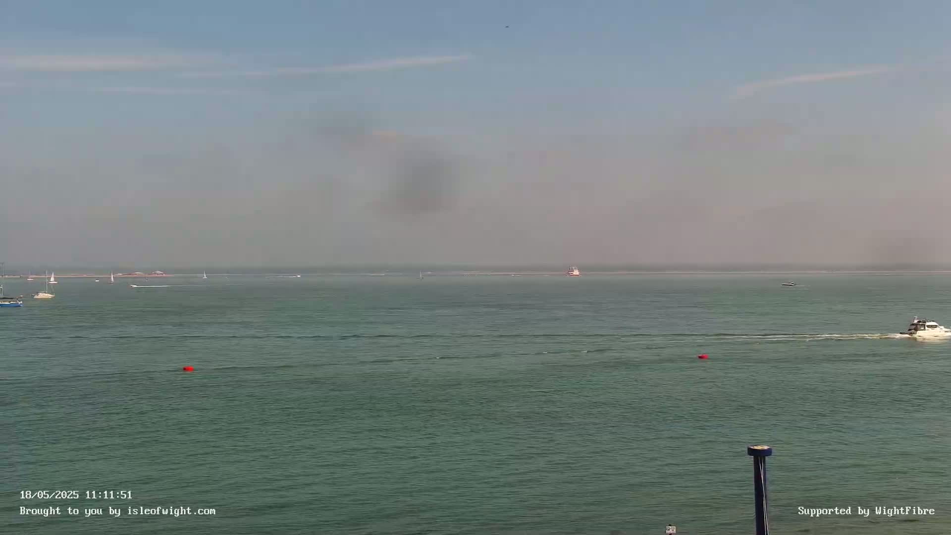 A hazy, calm sea with several boats and buoys visible, under a clear, light-blue sky.