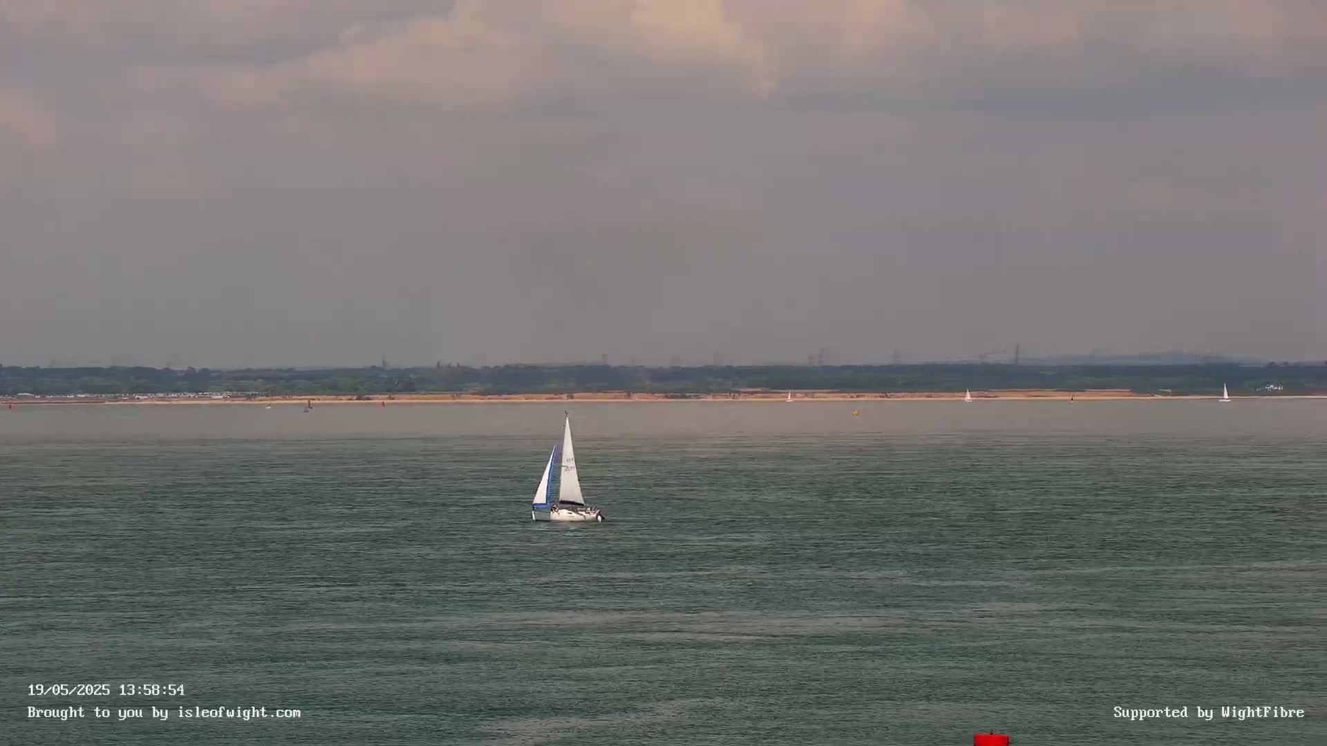 A sailboat is visible on calm, gray-green water under an overcast sky, with a distant shoreline in the background.