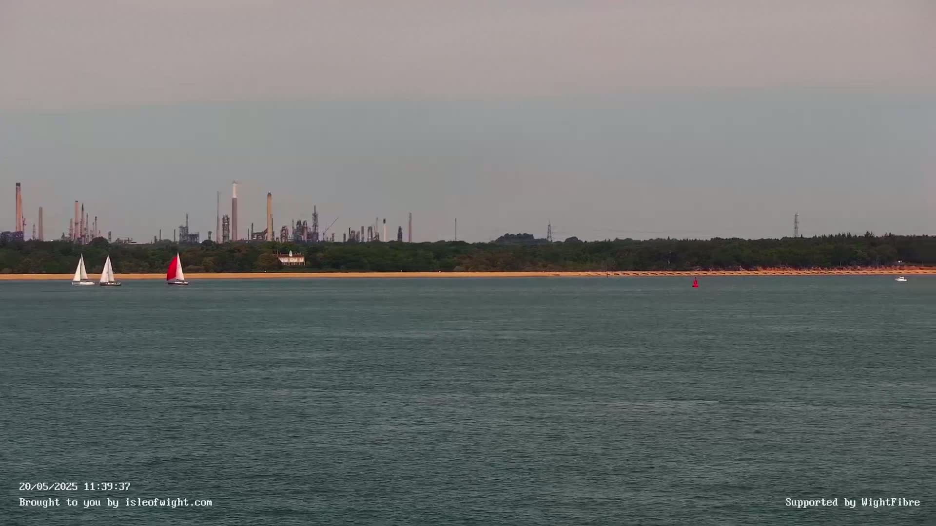 Three sailboats are visible on calm water in front of a sandy shore and an industrial refinery under a cloudy sky.