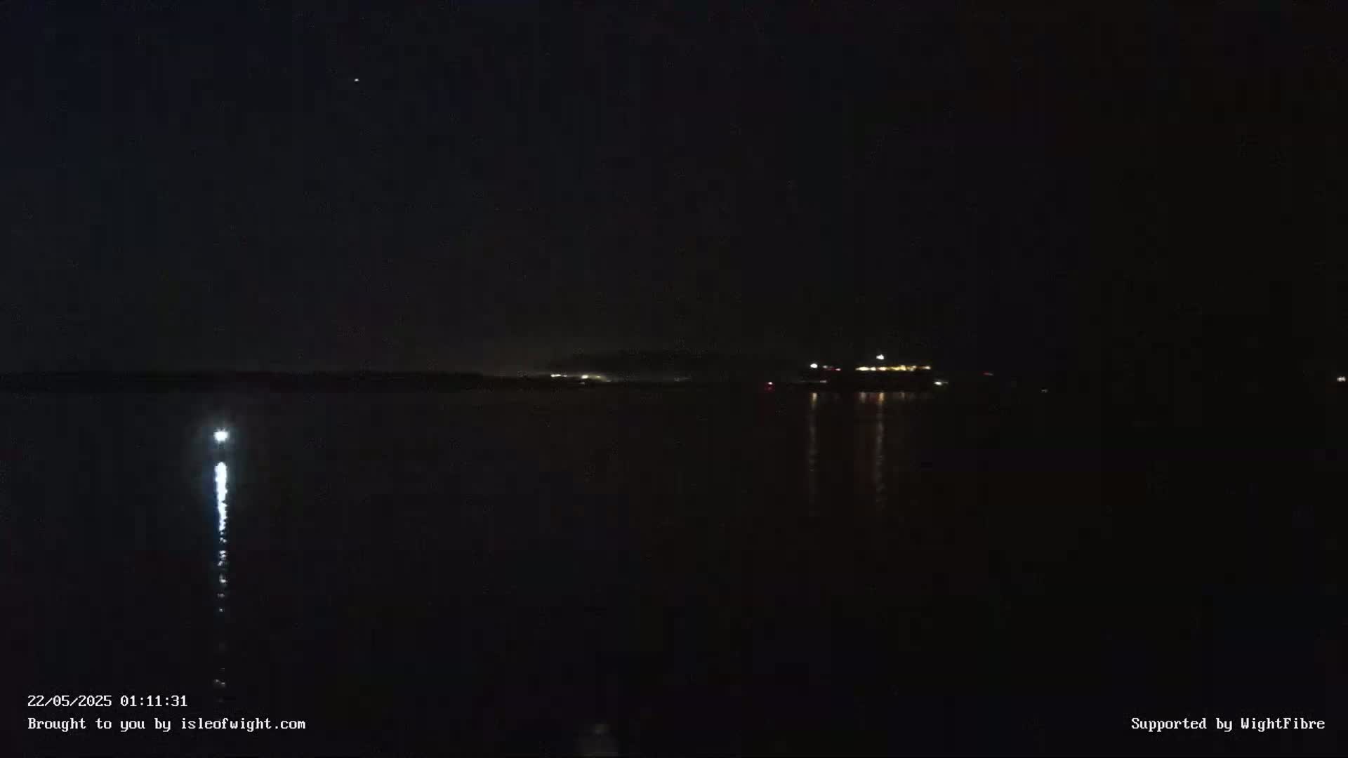 A dark nighttime scene shows a calm body of water reflecting a few distant lights under a clear, starry sky.