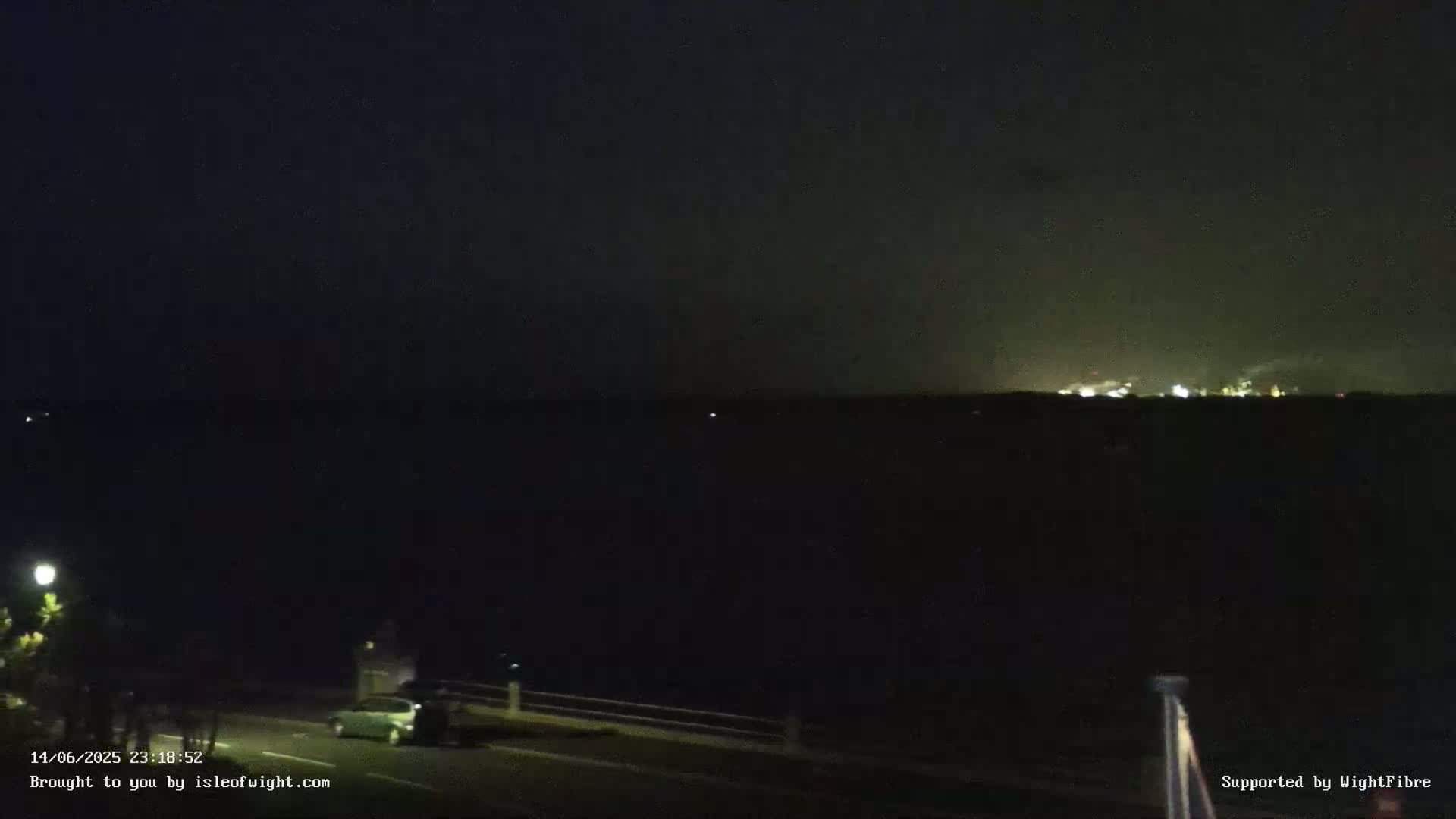 A dark nighttime scene shows a calm body of water reflecting a few distant lights under a clear, starry sky.