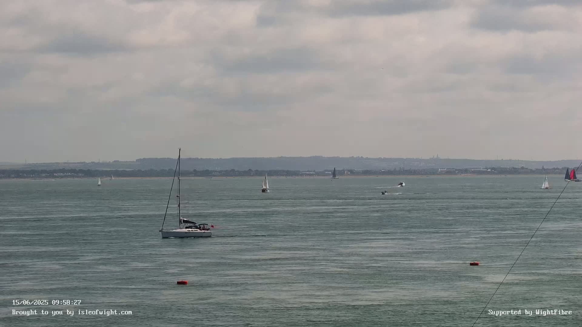 Several sailboats and motorboats are visible on a calm, gray day on a body of water with a distant shoreline.