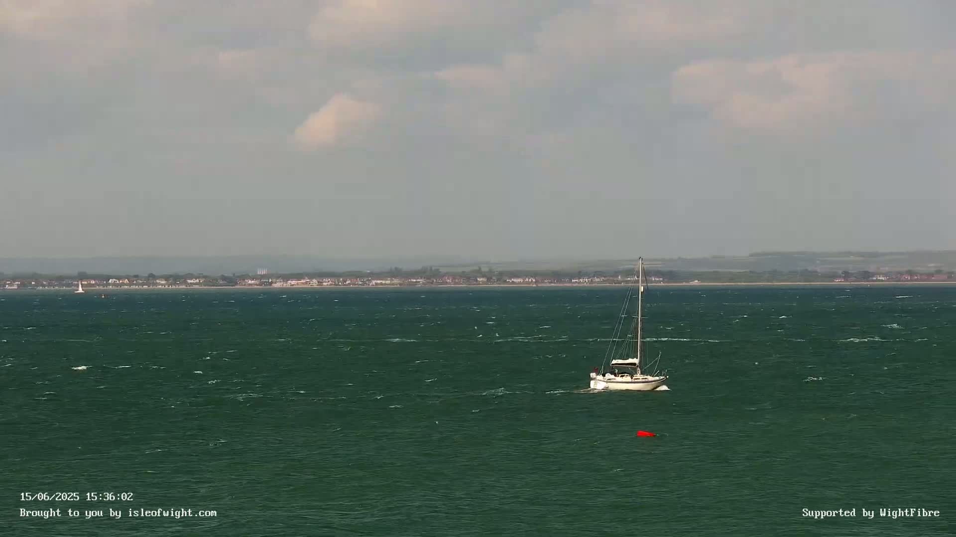 A sailboat is on a choppy, dark green sea under a mostly cloudy sky with a distant shoreline visible.