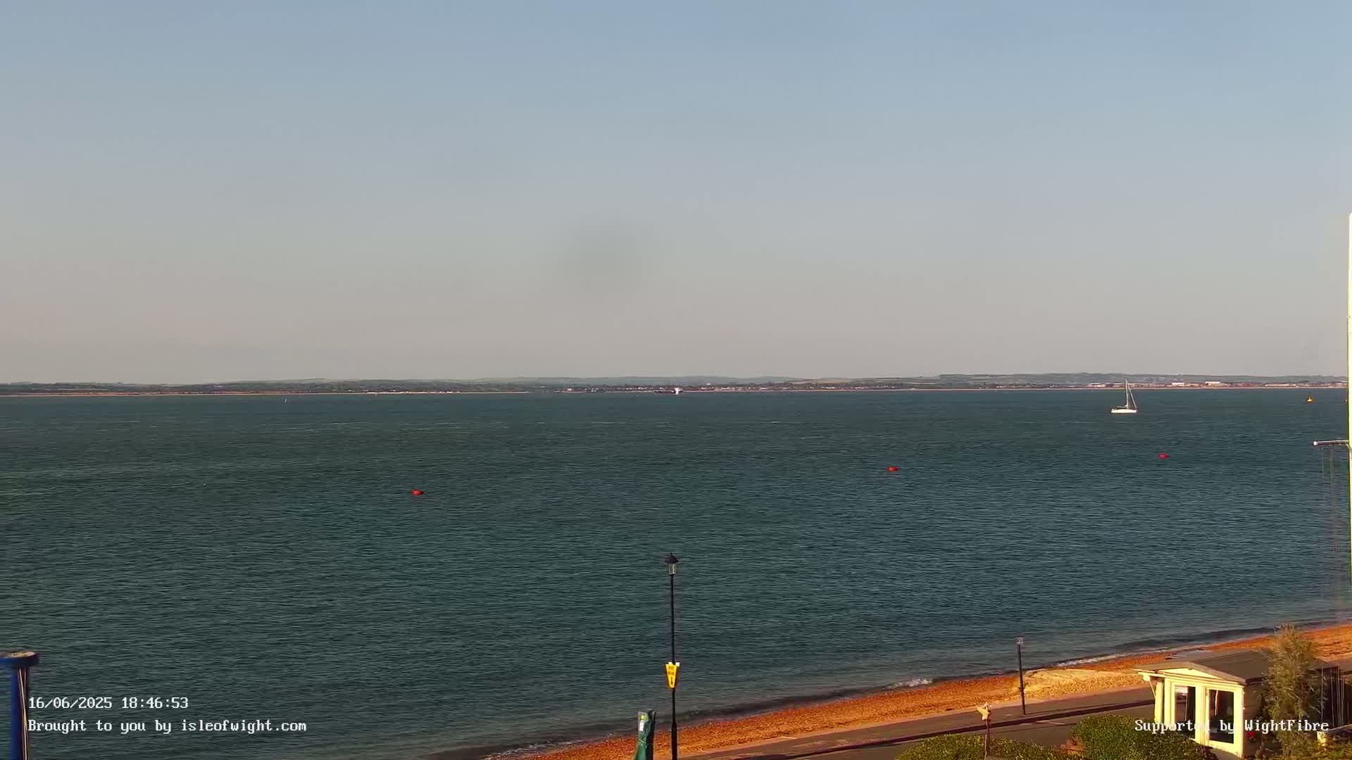 A calm body of water with a sailboat in the distance, a sandy beach, and a clear sky.