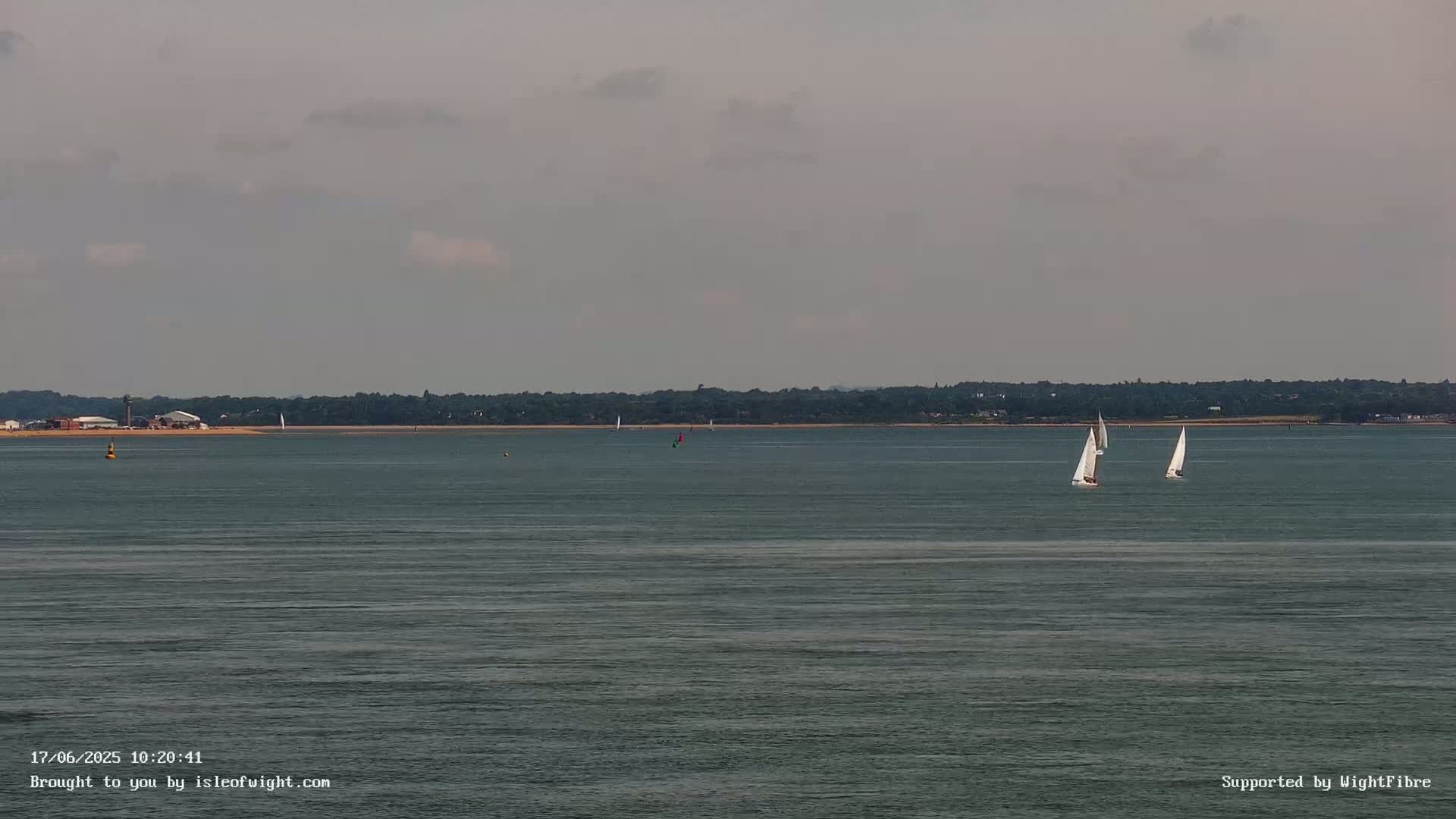 Two sailboats are visible on a calm, gray day on a body of water with a distant shoreline.