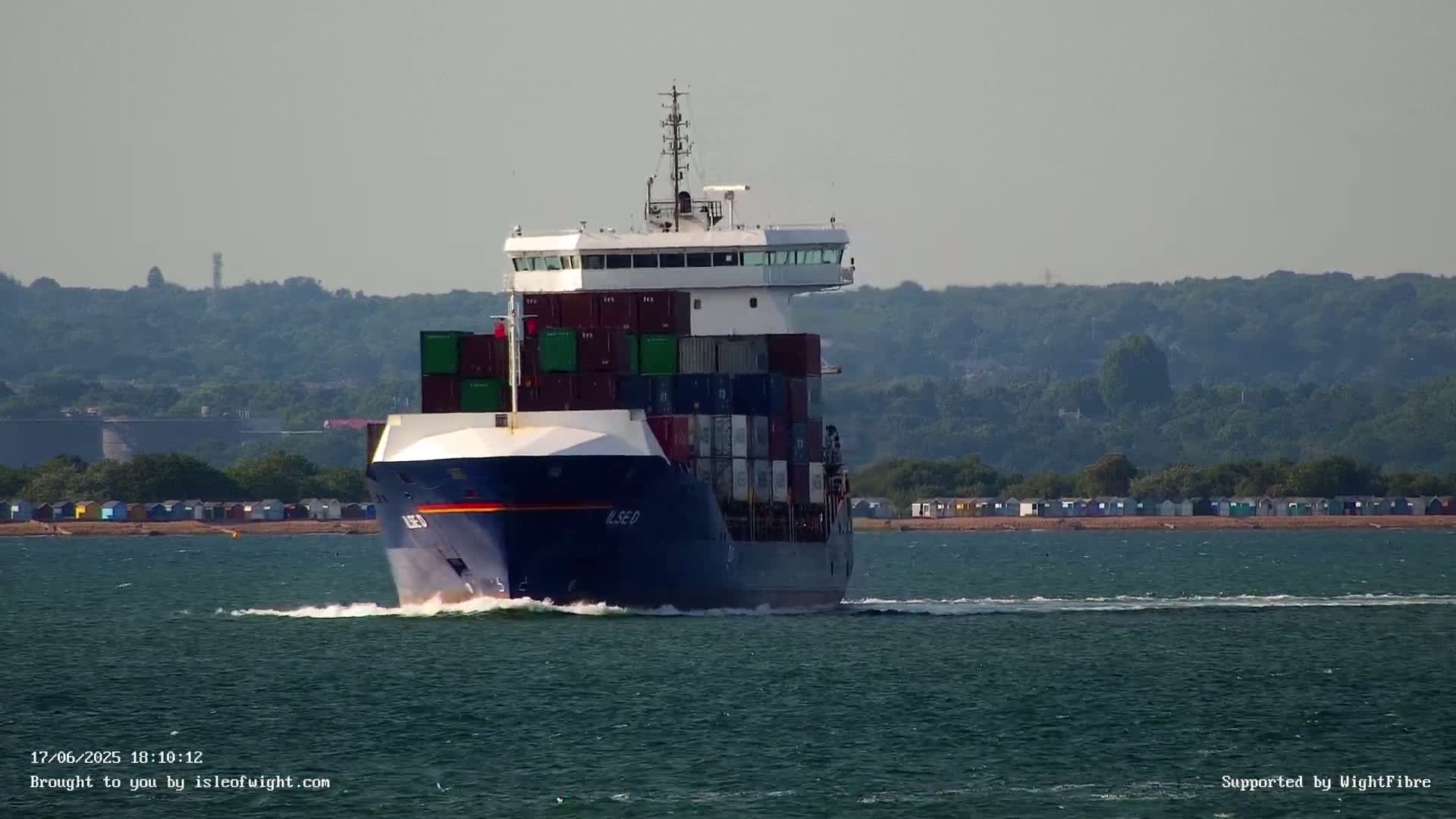 A container ship sails on calm water toward a shoreline with a row of colorful beach huts and a background of lush green hills.