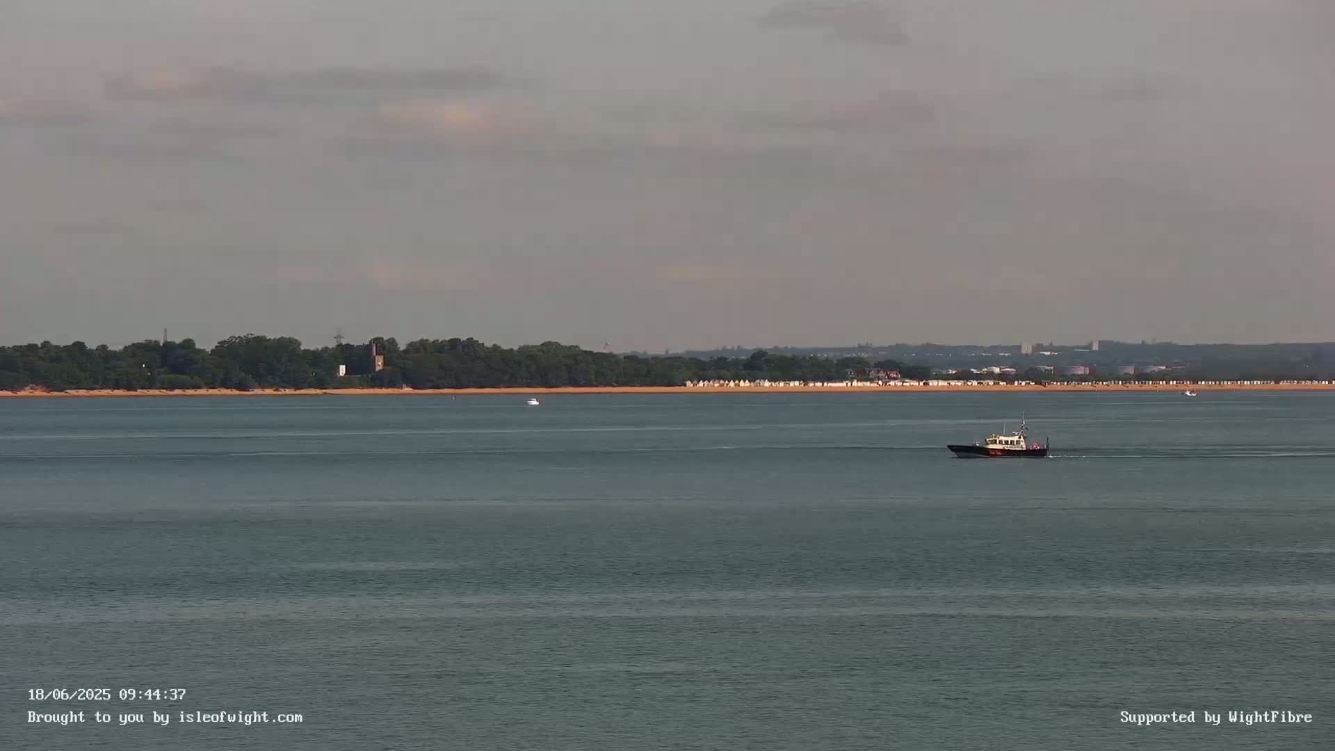 A small boat travels across calm, gray water toward a distant shoreline lined with trees and small structures under an overcast sky.