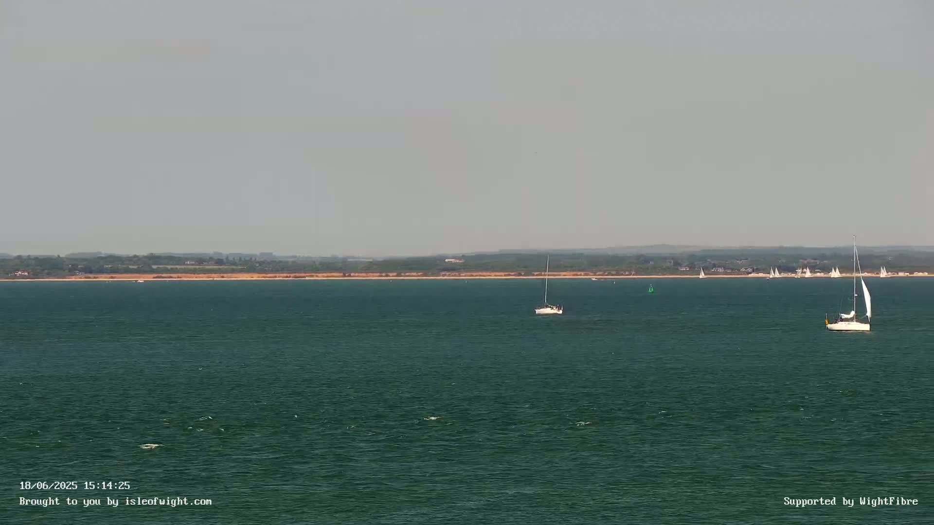A calm, hazy day shows a wide expanse of dark-green water with several sailboats, and a distant shoreline.