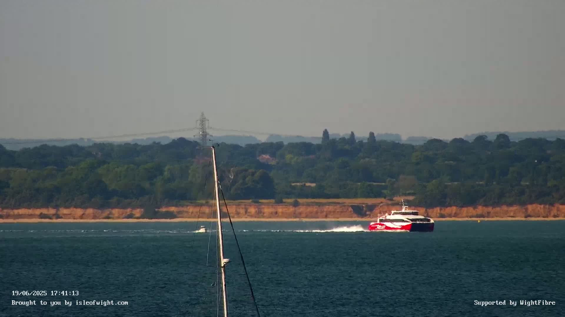A red and white ferry speeds across a calm body of water, past a smaller boat and towards a wooded shoreline under a hazy sky.