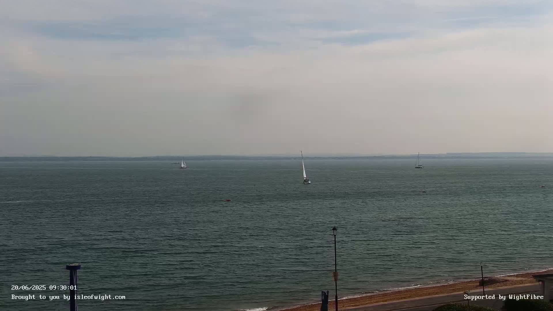 A hazy, calm sea with several sailboats visible in the distance, a sandy shore in the foreground.