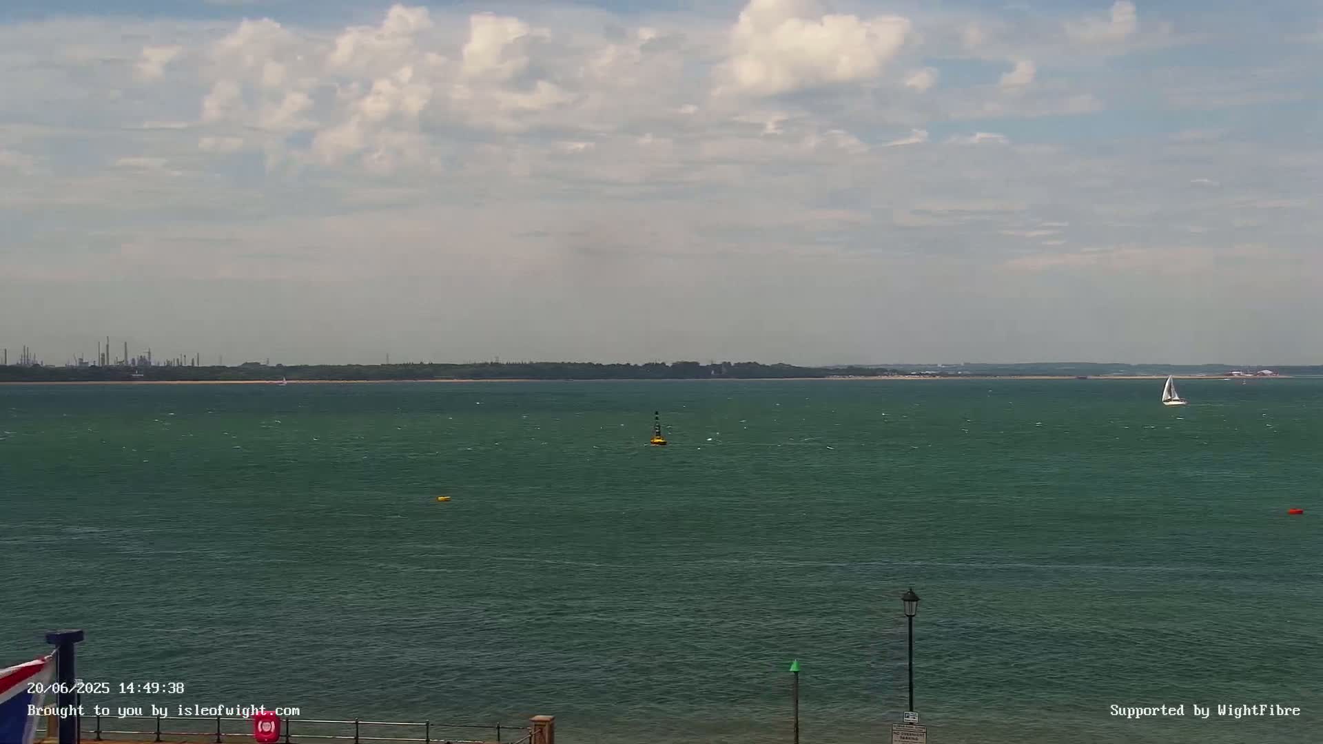 A partly cloudy day shows a wide expanse of dark-green water with a sailboat and a few small buoys, a distant shoreline with industrial structures, and a lamppost in the foreground.