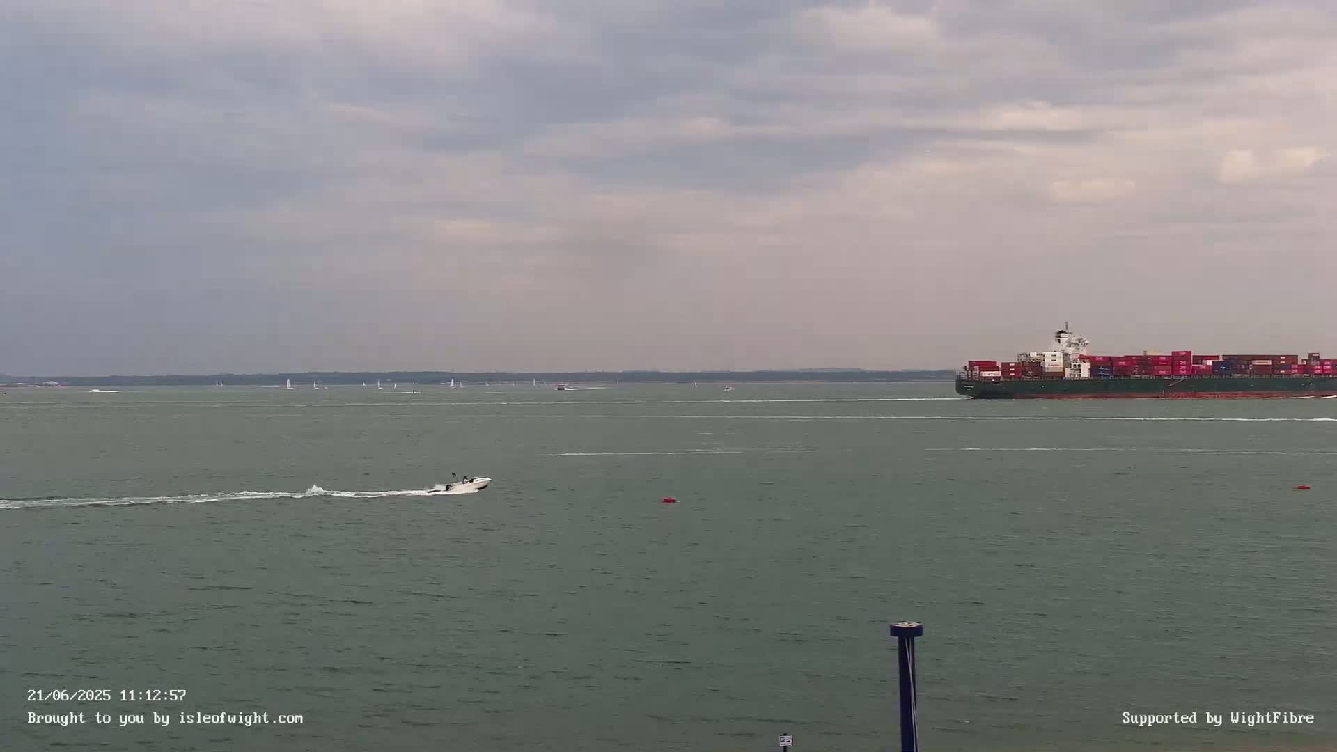 A large container ship and a small motorboat are visible on a calm, overcast sea with numerous sailboats in the distance.
