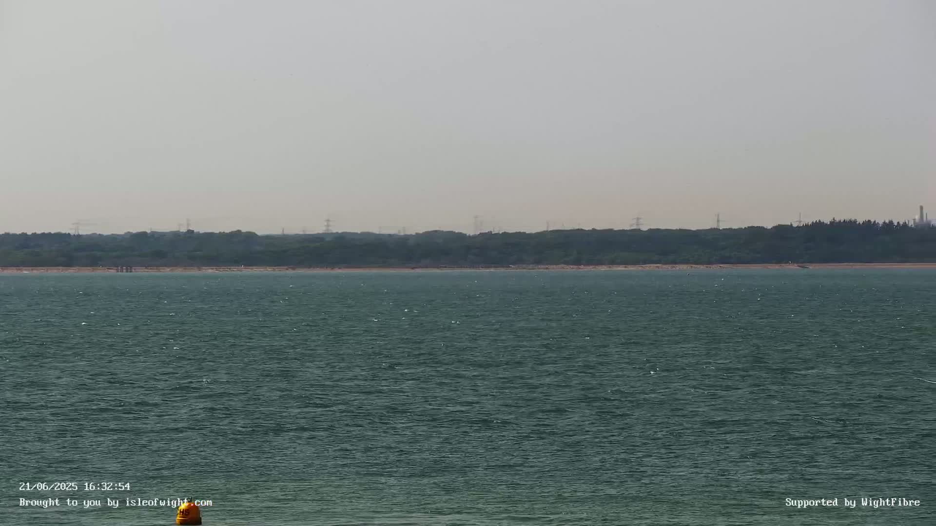A calm, dark-green body of water stretches to a hazy shoreline of trees and distant structures under a light-grey sky.