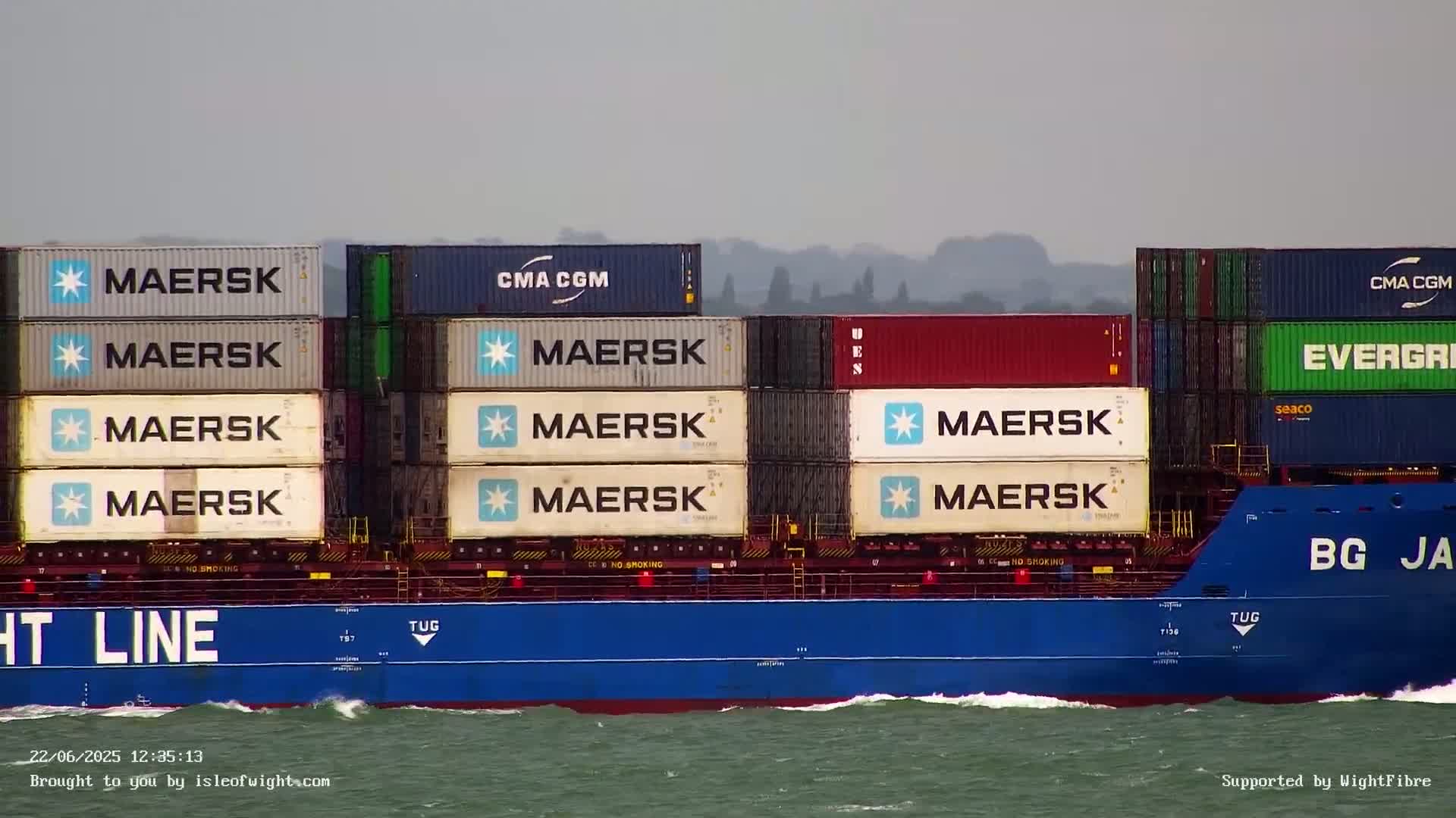 A large container ship, carrying numerous shipping containers, sails on a choppy sea under an overcast sky.