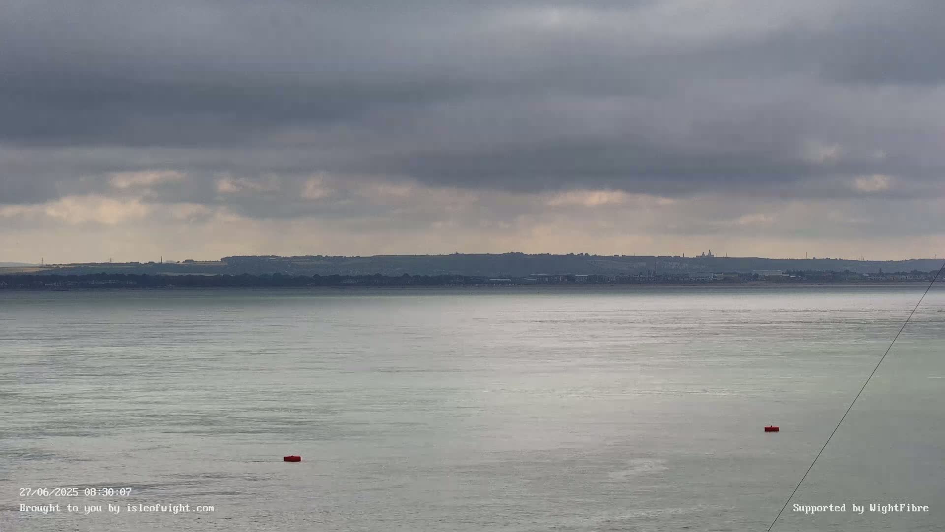 A calm body of water stretches to a distant shoreline under a cloudy sky, with two small red buoys visible on the water's surface.