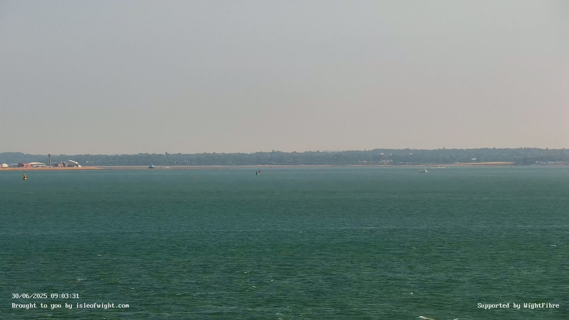 A hazy, calm sea stretches to a distant shoreline of low-lying land covered with vegetation.