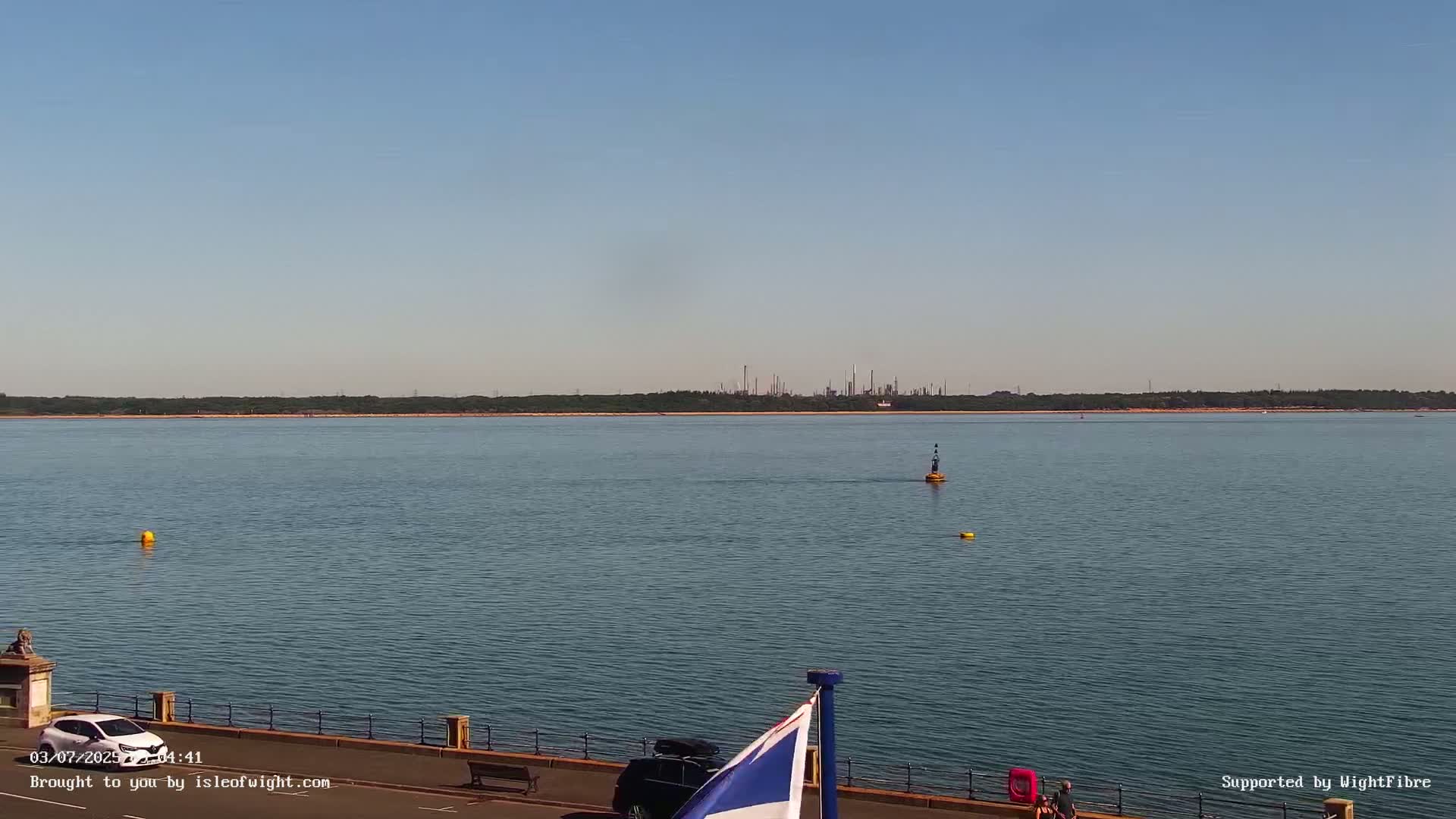 A calm body of water stretches to a distant shoreline featuring an industrial complex under a clear, bright sky.