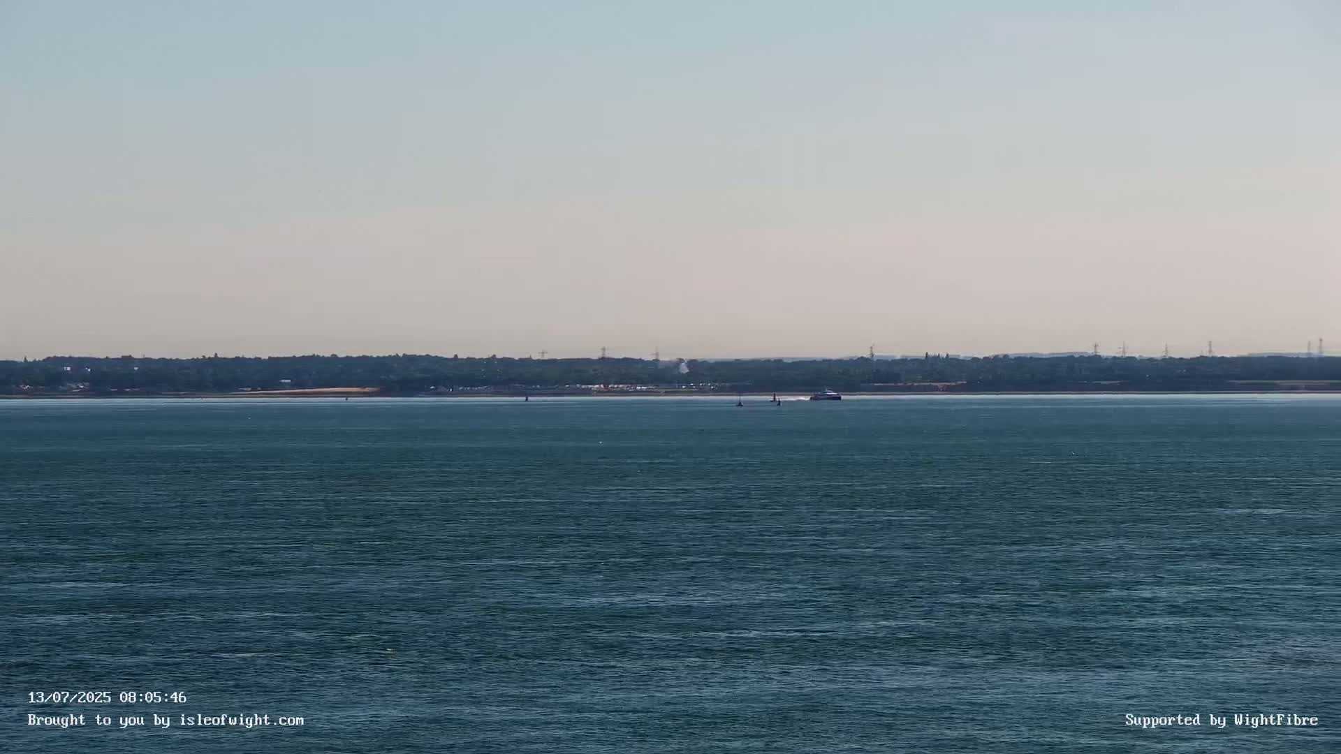 A calm body of water stretches to a distant shoreline of trees and structures under a clear sky.