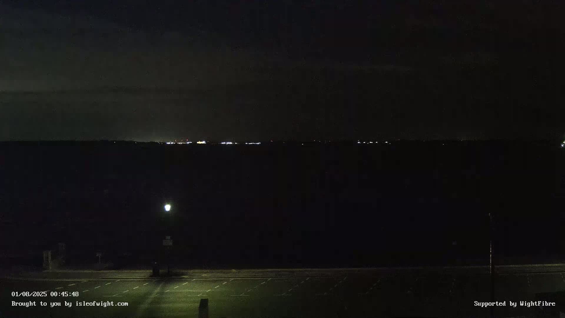 A dark, nighttime scene shows a distant line of lights across a body of water, a lamppost in the foreground illuminating an empty parking lot.