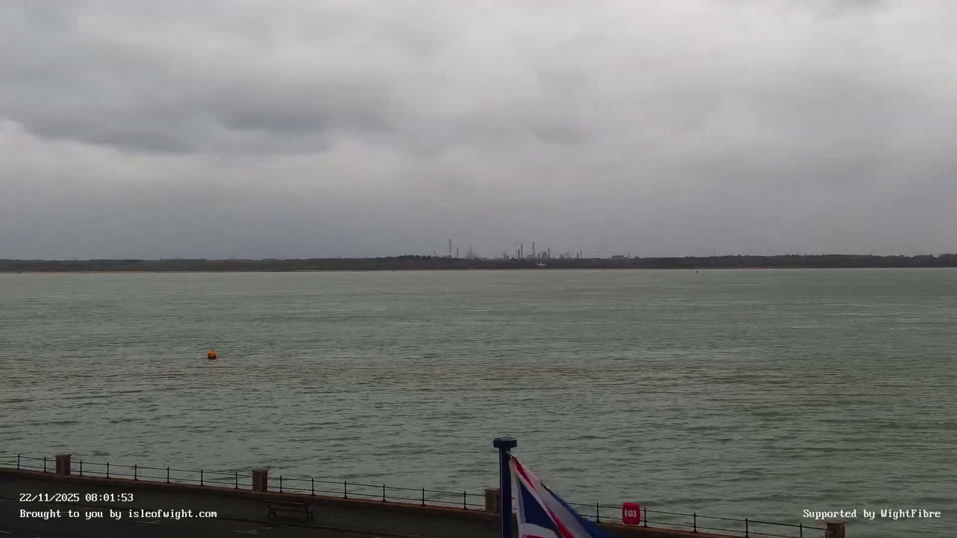 Under a heavy overcast sky, a wide body of water with a single yellow buoy stretches towards a distant forested shoreline featuring an industrial complex, all viewed from behind a dark promenade railing.