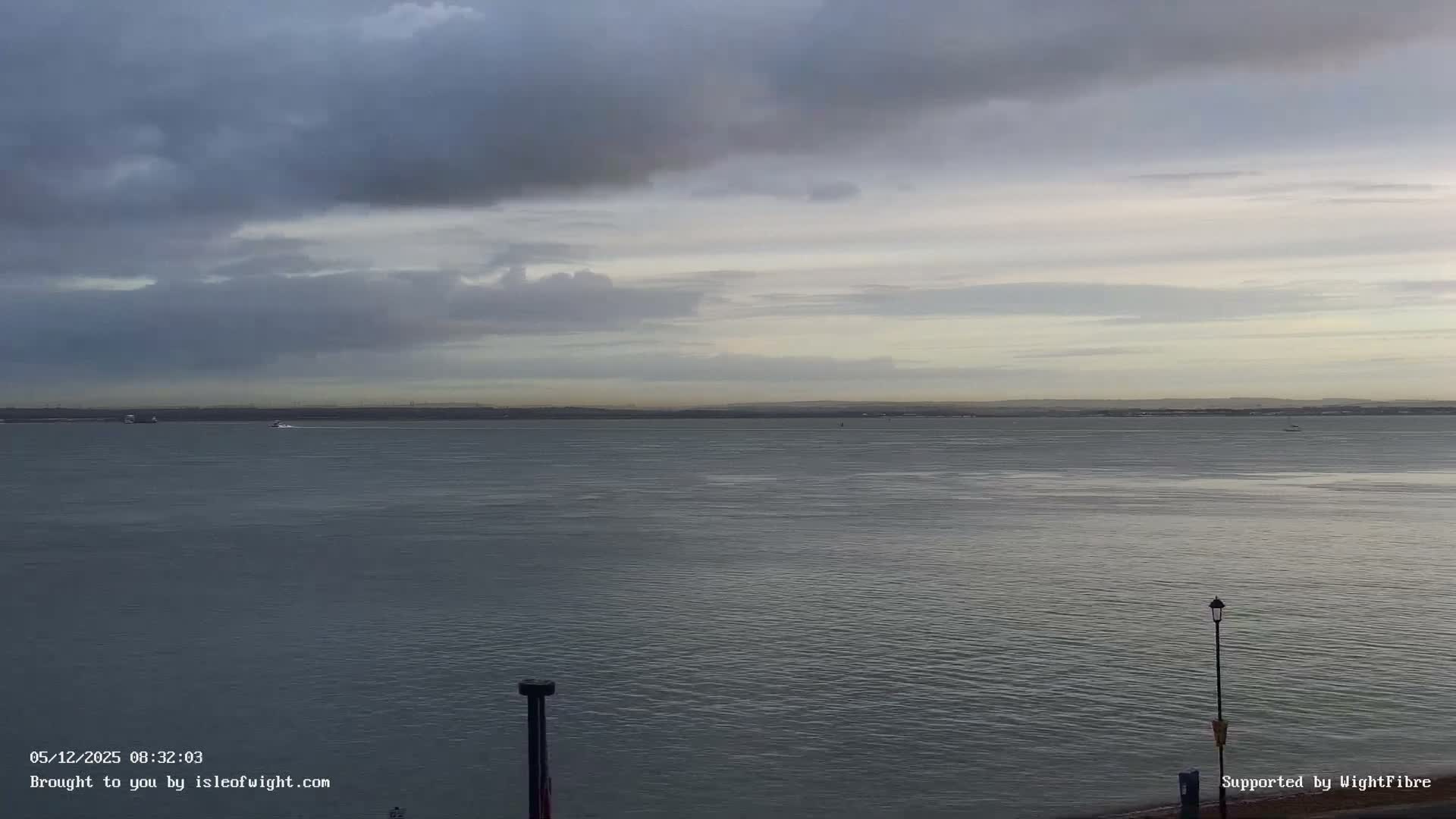 Under a heavily overcast and grey sky, a calm body of water stretches towards a hazy distant shoreline, dotted with a few boats and partially framed by dark structures in the foreground.