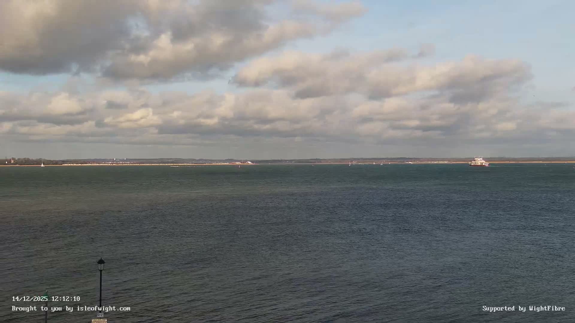 Under a heavily overcast and grey sky, a calm body of water stretches towards a hazy distant shoreline, dotted with a few boats and partially framed by dark structures in the foreground.
