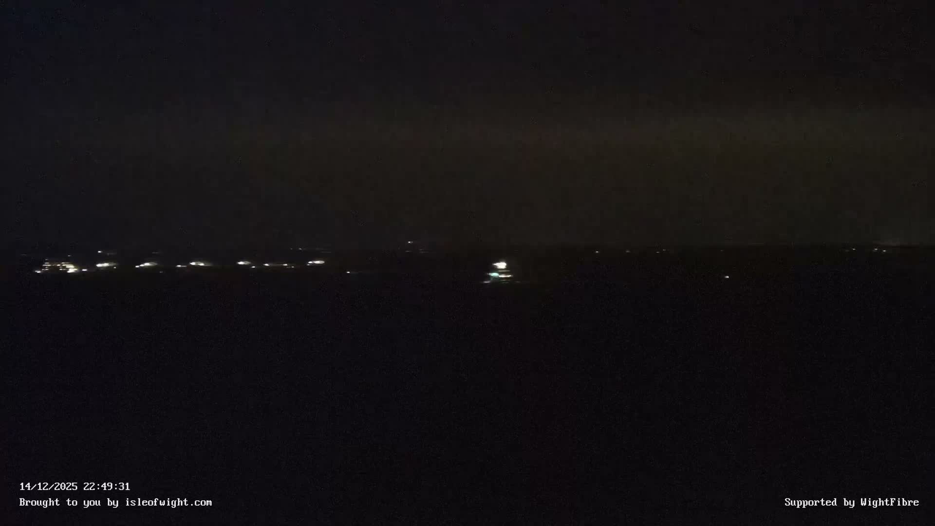 Under a heavily overcast and grey sky, a calm body of water stretches towards a hazy distant shoreline, dotted with a few boats and partially framed by dark structures in the foreground.
